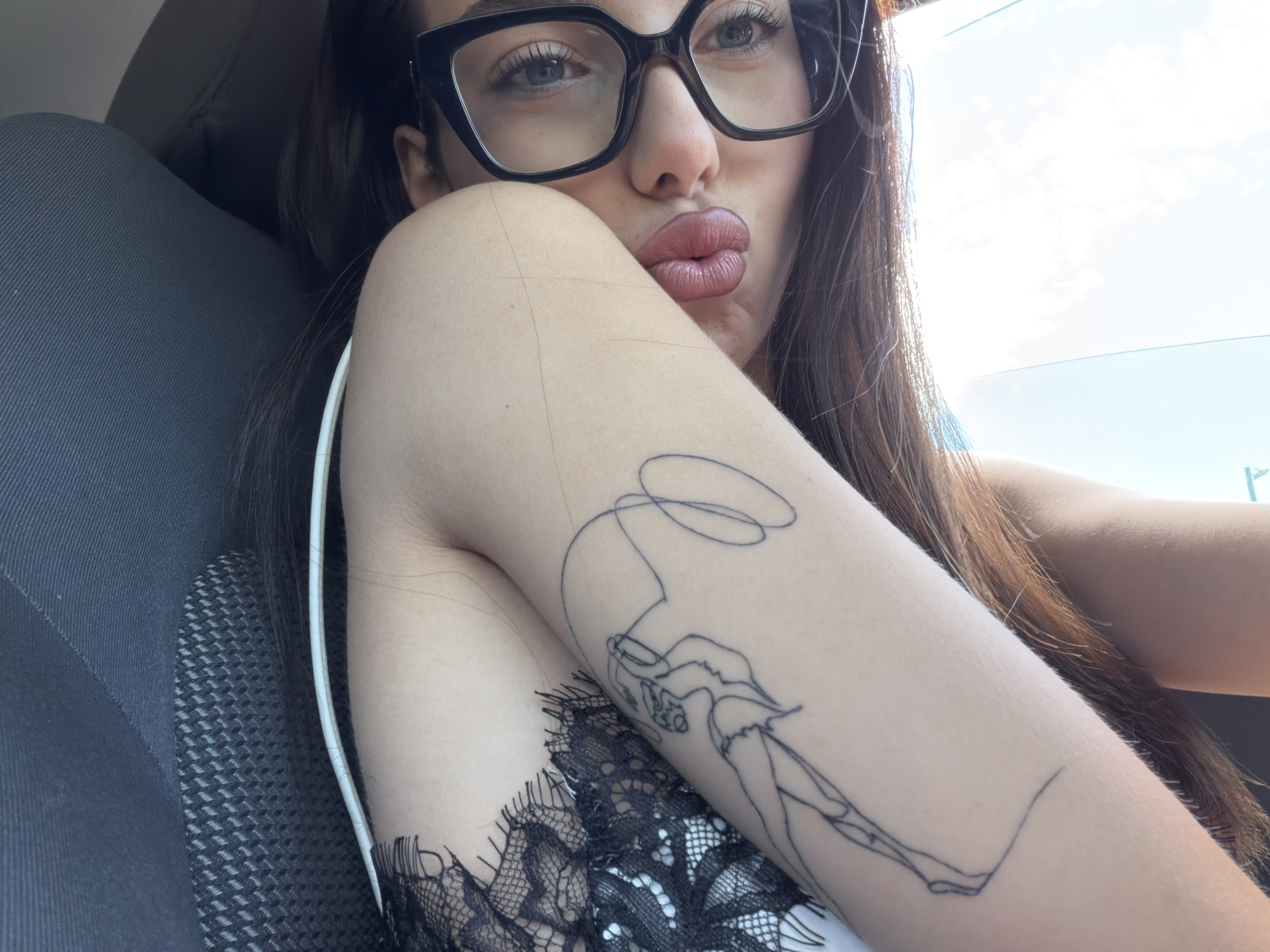 A young woman with long brown hair and glasses is sitting in the driver's seat of a car. She is wearing a black lace top and has a tattoo of a dancer on her right arm. The woman is looking at the camera and pouting her lips. The car appears to be parked in an outdoor location with the sky visible through the window.