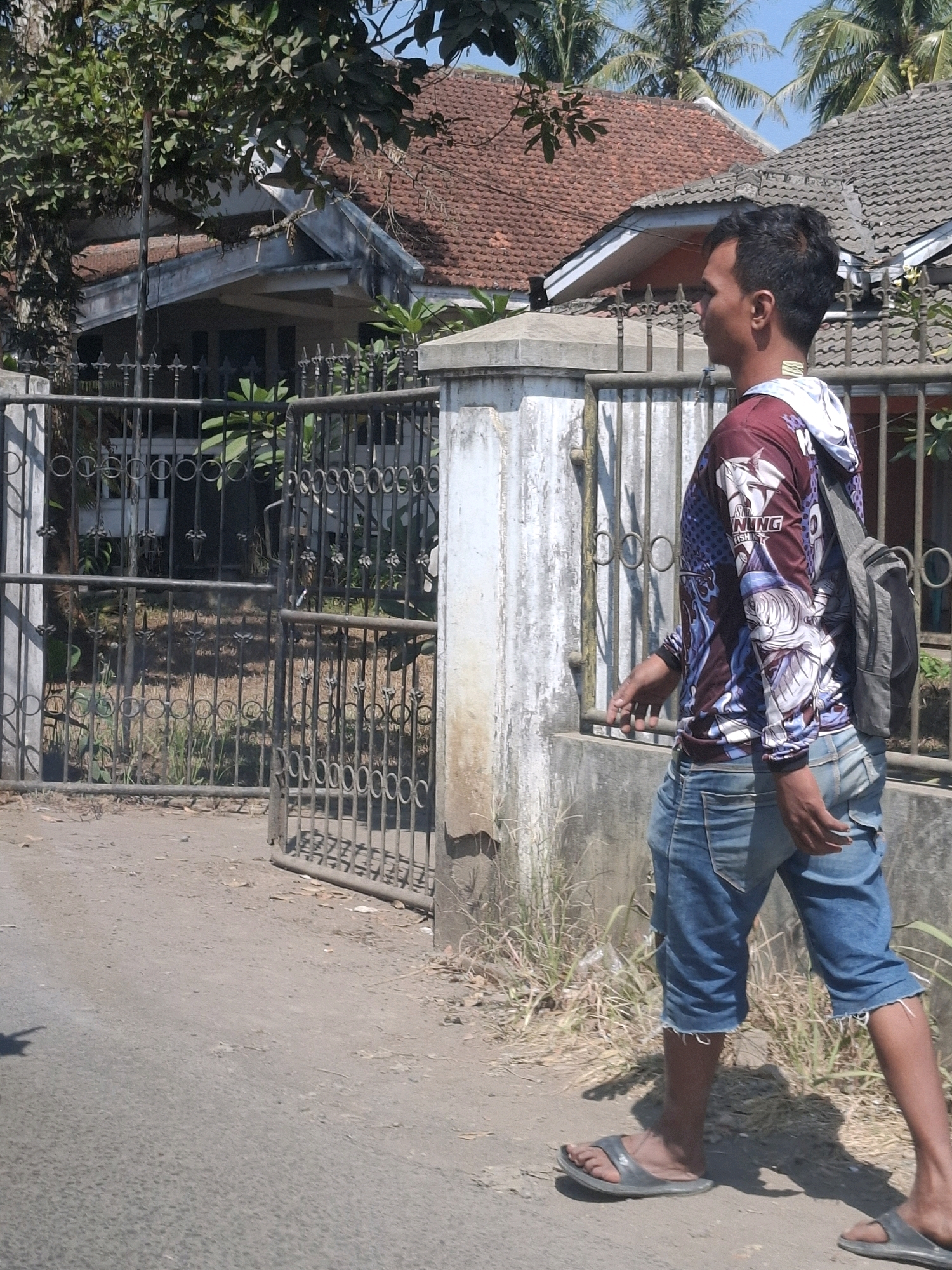 A man wearing a long-sleeved shirt with a graphic print and blue denim shorts is walking along a dirt road next to a metal fence. He has a grey backpack slung over his right shoulder and is wearing grey flip-flops. Behind the fence, a house with a red tile roof is visible, surrounded by trees and vegetation. The scene appears to be in a rural or suburban area, possibly in Cisaat, Indonesia, given the architectural style and vegetation. The lighting suggests it is daytime.
