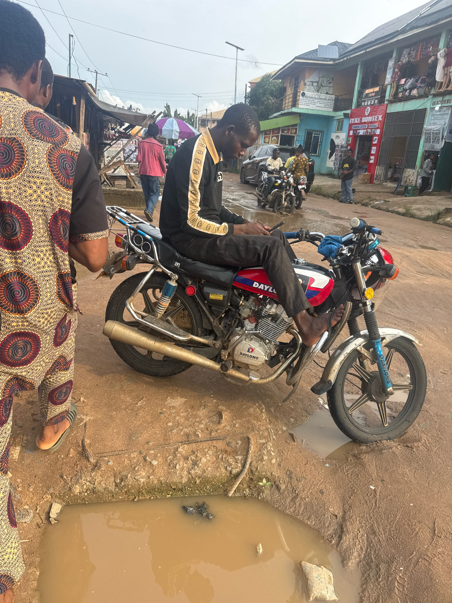 In Ihiagwa, Nigeria, on a muddy dirt road, a young Black man sits astride a red, white, and blue Daylong motorcycle. He is dressed in black pants with a gold and black striped jacket and is looking at his phone. Beside him, to the left, a person wearing a colorful patterned dashiki and black pants faces away from the camera. In the background, a row of shops with corrugated metal roofs line the street. One shop has a red banner with white lettering that reads "AYKE BONITEX COMMUNICATION VENTURES." Several other motorcycles and a car are visible further down the road, and a large muddy puddle occupies the foreground. The sky is overcast.