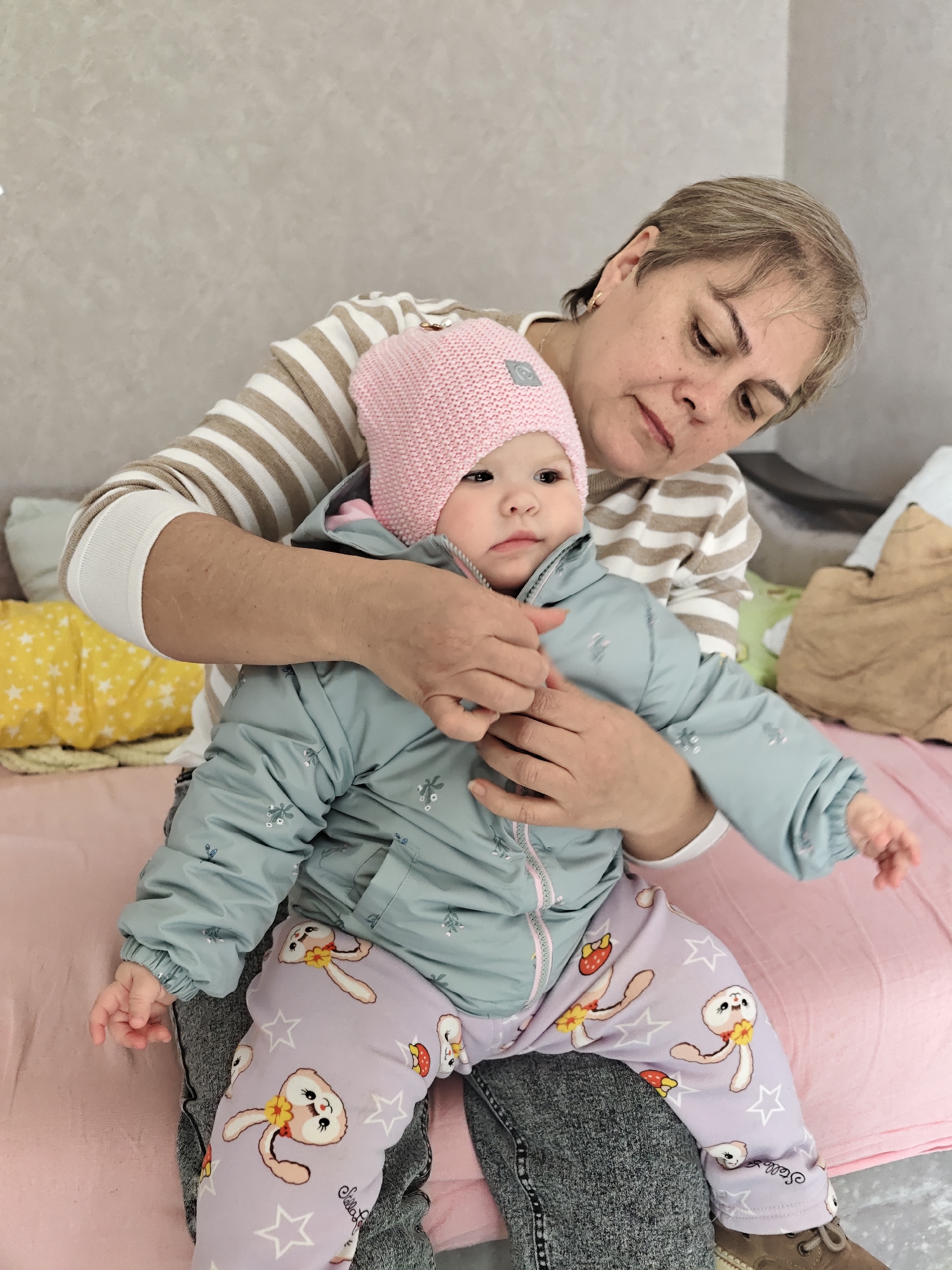 A woman is holding a young child while seated indoors on a pink bed. The woman is fair-skinned with short, graying hair and is wearing a horizontally striped, long-sleeved shirt. The child, who is fair-skinned and wearing a pink knit hat, is dressed in a light blue puffer jacket and patterned pants featuring bunny illustrations and stars. The woman is holding the child's hands, appearing to be assisting or comforting the child. In the background, there's a gray wall, a yellow star-patterned pillow, and a brown object. It is not possible to confirm the location is in the city of Chaikovsky, Russia.