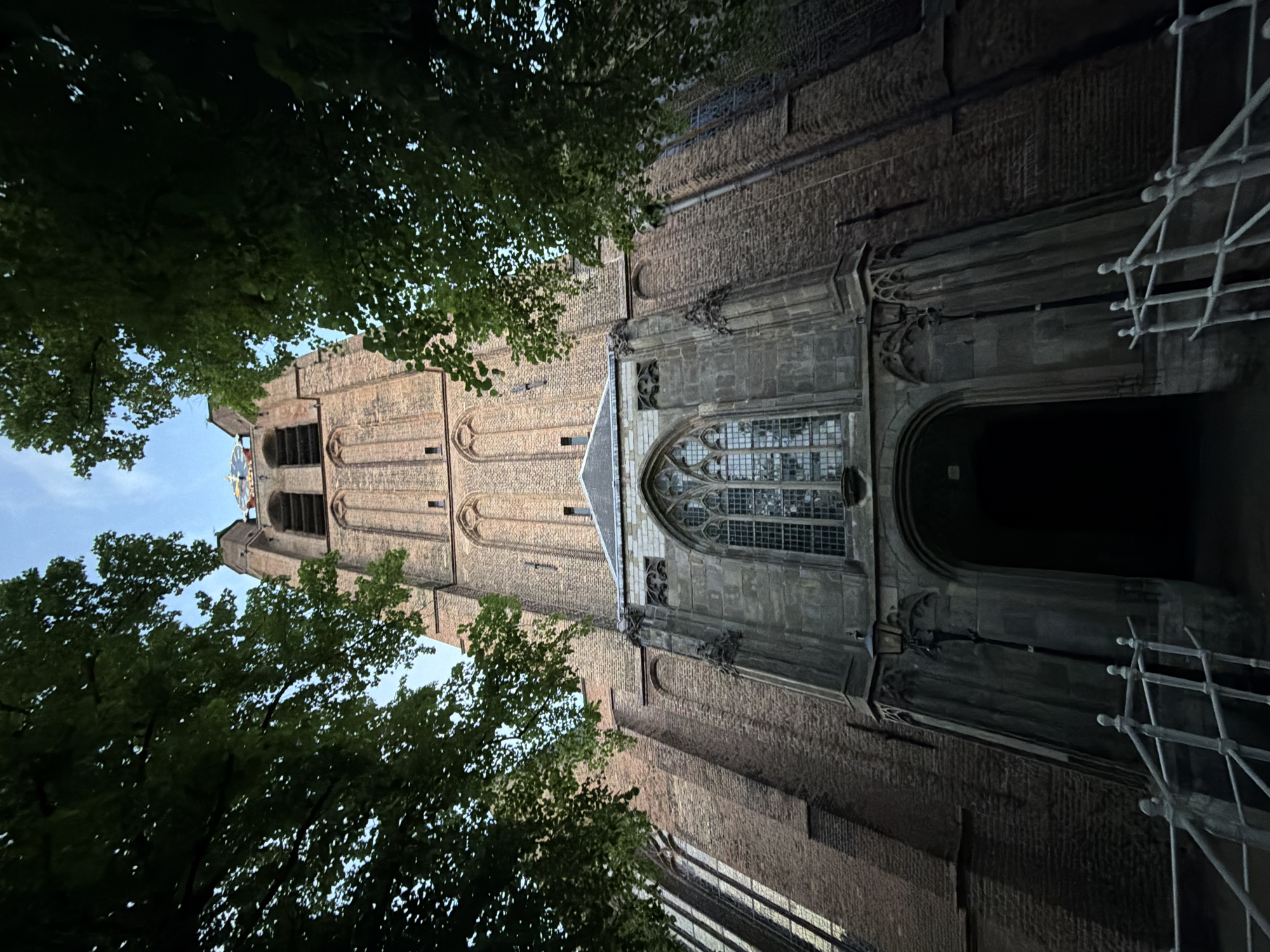 The image captures a low-angle view of the Oude Kerk (Old Church) in Delft, Netherlands. The church's brick facade dominates the scene, with a tall, multi-tiered tower rising towards the sky. A large Gothic window is visible on the side of the church, adjacent to a dark entranceway. Scaffolding stands near the entrance, suggesting ongoing restoration or maintenance work. Green foliage from trees partially obscures the view of the tower and frames the left side of the image, contrasting with the church's brick. The sky is visible in the upper left corner.