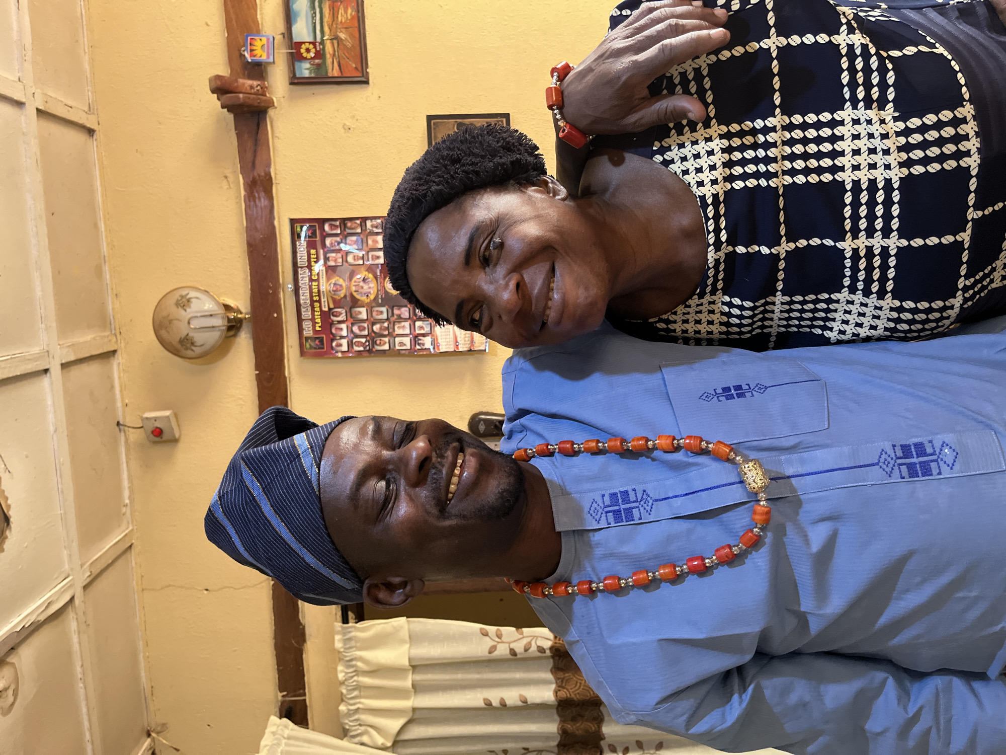 Two people are posing for a photo indoors. A man with dark skin, a dark beard, and a striped blue hat is lying down, smiling, and wearing a light blue long-sleeved shirt with blue embroidery and a beaded necklace. A woman with dark skin and short black hair is leaning over him, smiling, and wearing a dark blue and white patterned dress with a beaded bracelet. The background includes a yellow wall with a light fixture, various pictures hanging, and a white door. The location is Rafin Bauna, Nigeria.