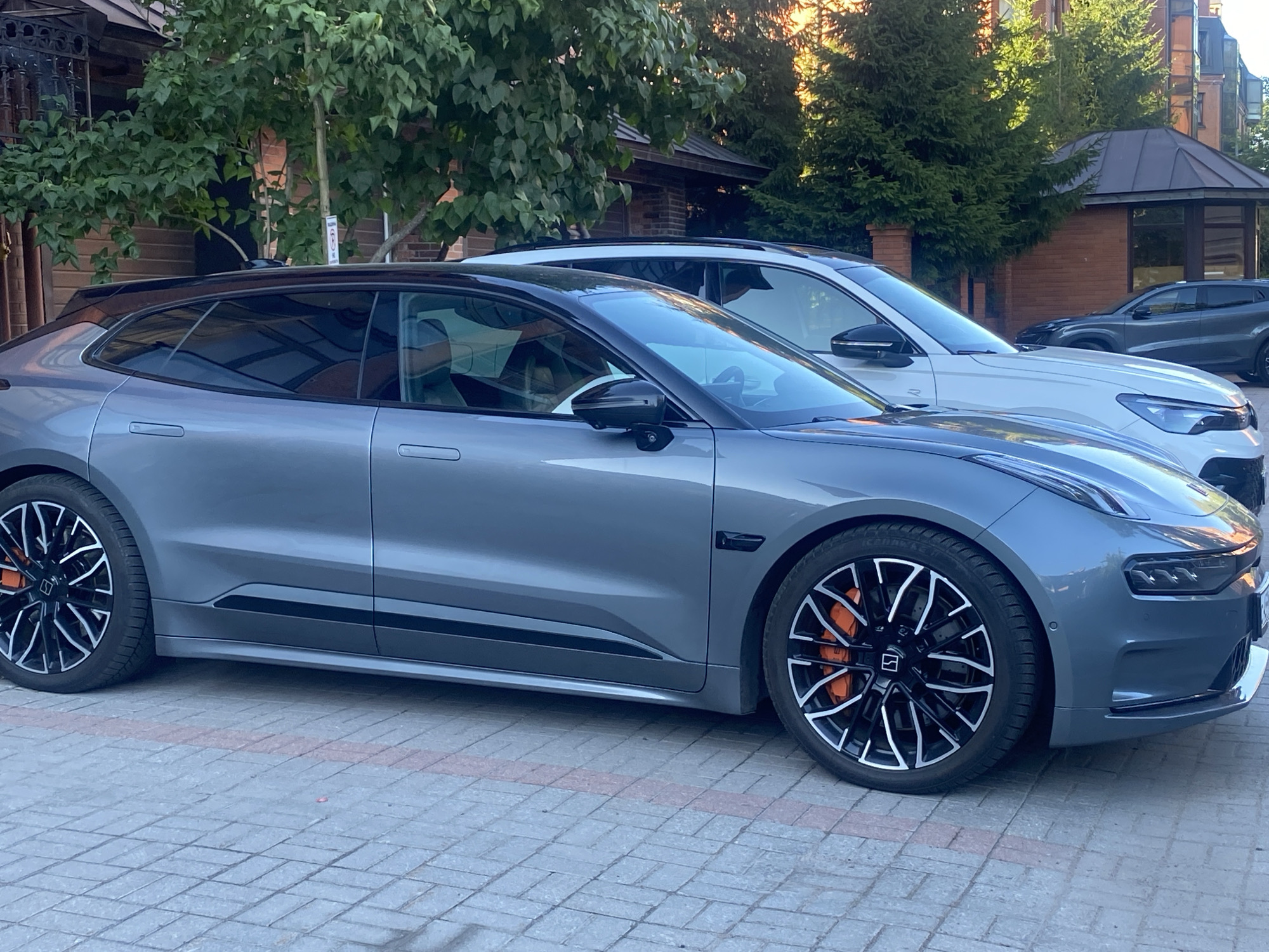 Two modern cars are parked on a paved area. The car in the foreground is a grey electric hatchback with black trim and distinctive black and silver wheels with orange brake calipers. The car in the background is a white SUV. Behind the cars, there is a brick building with large windows and a brown-tiled roof, surrounded by lush green trees and evergreen shrubs. The overall setting suggests an upscale urban or suburban environment, possibly a private residence or a commercial establishment in Saint Petersburg, Russia. The lighting indicates it is daytime, with sunlight casting shadows and highlighting the vehicles and foliage. There are no people visible in the image.