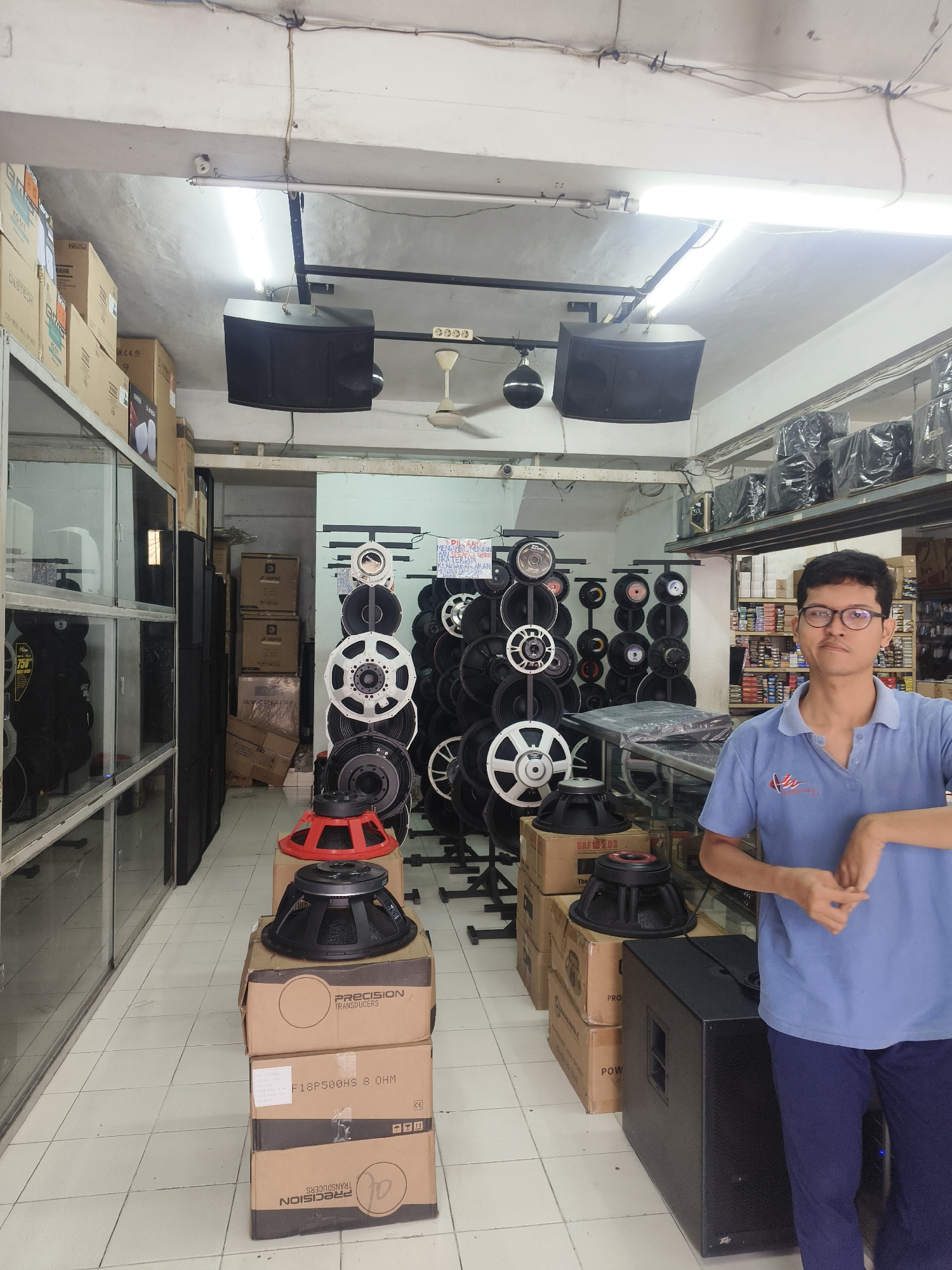 The image depicts the interior of an audio equipment store, likely located in Denpasar, Indonesia, characterized by numerous speakers and audio components. In the foreground, a young man wearing glasses, a blue polo shirt with a logo, and dark pants stands facing the camera. He is positioned to the right of the frame, with his hands clasped in front of him.
The store environment is filled with various audio equipment. Stacked on the floor are several cardboard boxes, some labeled with brand names like "PRECISION TRANSDUCERS" and product codes such as "F18P500HS 8 OHM". On top of these boxes are several large speaker drivers, including one with a distinctive red basket. To the left of the man and boxes, there is a display unit with glass shelves containing more audio equipment, and further to the left, a tall, black speaker cabinet is visible.
The background reveals a wall lined with numerous speaker drivers of different sizes, mounted on racks. Above, speakers are suspended from the ceiling, and fluorescent lights illuminate the space. The floor is tiled with a light-colored pattern. Shelving units filled with more equipment and boxes are visible further into the store. The overall impression is of a well-stocked retail space dedicated to audio systems.