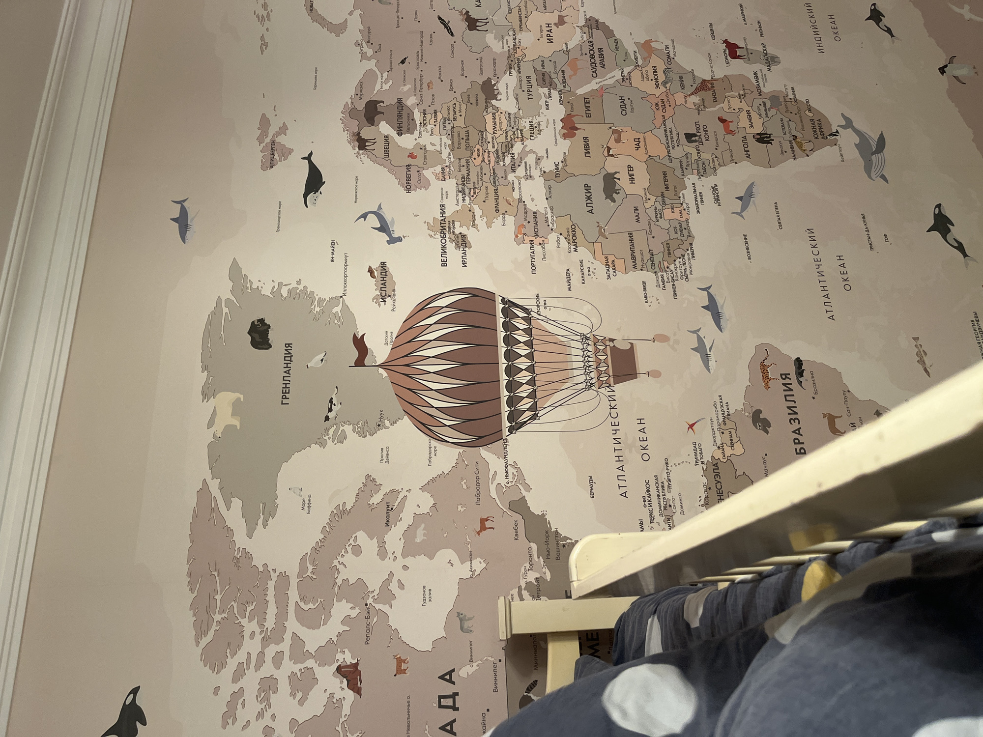The image shows a world map wallpaper on a wall, partially obscured by a crib. The map includes continents, countries, and oceans labeled in Russian. A hot air balloon illustration is centered on the North Atlantic Ocean. Animal illustrations, such as whales and sharks, are scattered across the oceans. The crib, made of light wood, is positioned in front of the lower right portion of the wallpaper, with blue bedding visible inside. The overall lighting suggests an interior setting. The location of the city of Kaspiysk, Russia is not visible on the map, and therefore, not visible in the image.
