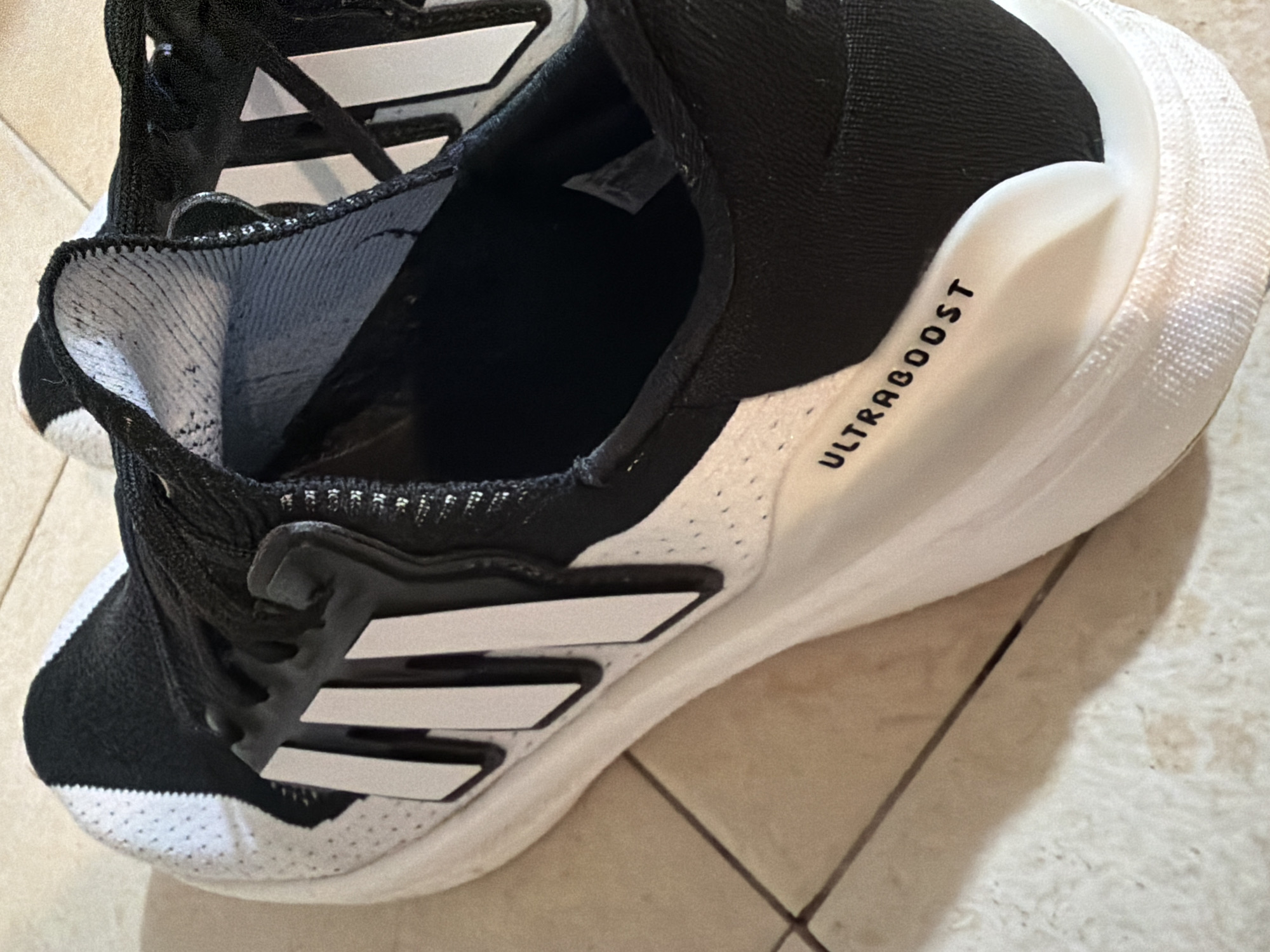 This is a close-up, eye-level shot of a black and white Adidas Ultraboost running shoe, resting on a tiled floor in Even Yehuda, Israel. The shoe is positioned on its side, with the inner sole visible. The Adidas logo, consisting of three white stripes on a black background, is prominently displayed on the side of the shoe. The word "ULTRABOOST" is printed in black lettering on the white midsole. The tiled floor, a neutral beige color with darker grout lines, serves as the backdrop. The lighting is direct and casts a shadow from the shoe onto the floor.