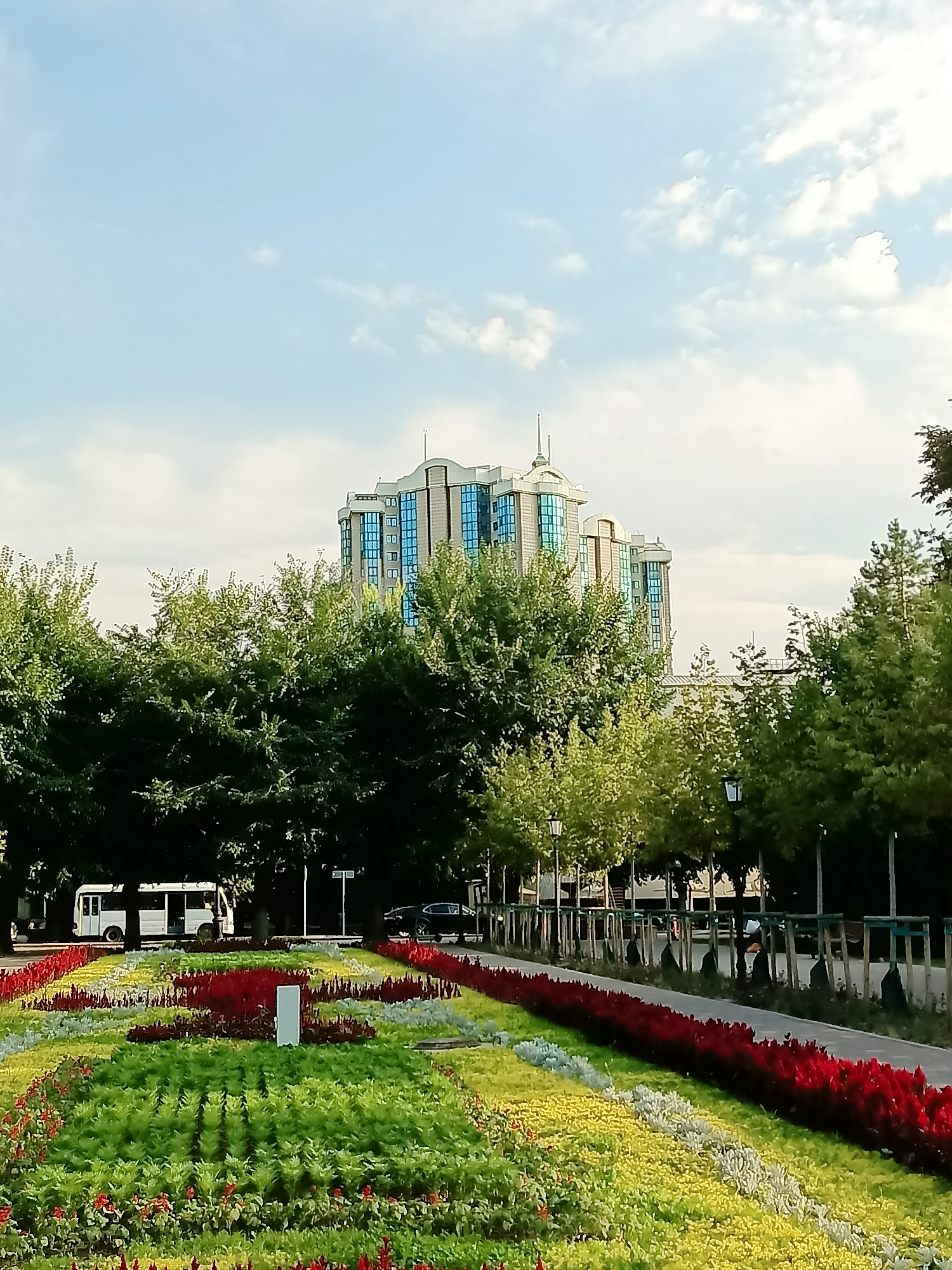 This image depicts a well-maintained public park in Almaty, Kazakhstan, likely located in a central or administrative area given the presence of a modern, multi-story building in the background. The building features a distinctive design with a stepped facade and numerous blue glass windows, suggesting it could be an office building or a government facility.
In the foreground, a vibrant flowerbed displays a colorful arrangement of red, yellow, and green plants, creating a strong visual contrast with the lush green trees that form a middle ground. The trees are mature and full, providing shade and framing the scene.
To the left, a white bus is parked on a paved road that runs parallel to the flowerbed. A dark-colored car is visible further down the road, indicating vehicular traffic. The road is lined with trees and lampposts, characteristic of a city park or boulevard.
The sky is partly cloudy with a bright blue hue, suggesting a pleasant day. The overall impression is one of an urban oasis, a harmonious blend of nature and modern architecture. The precise location within Almaty is not discernible from the image alone, but the architectural style of the building and the general landscaping are consistent with elements found in the city.