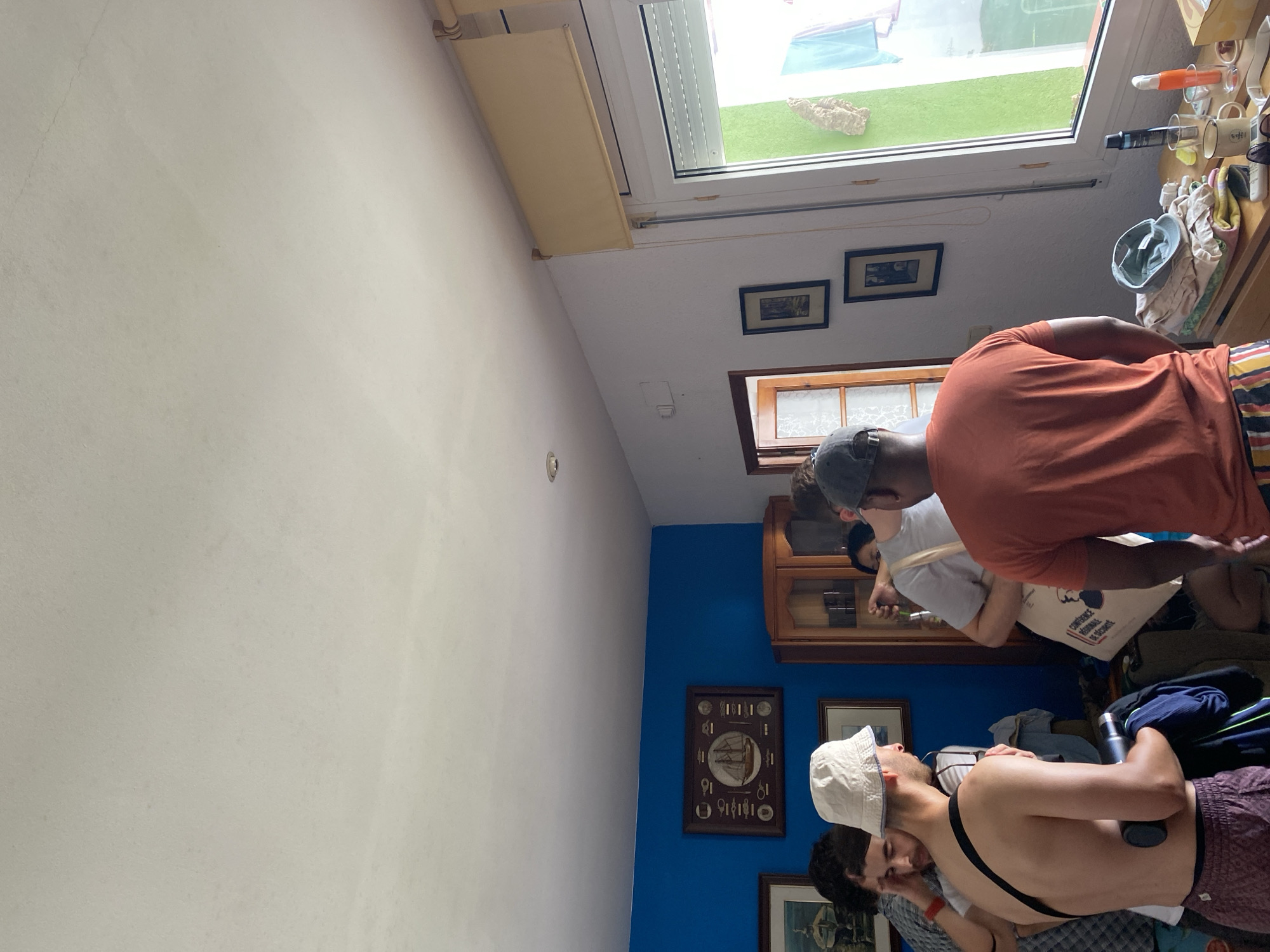 The image depicts an indoor scene with several individuals gathered in what appears to be a living room or common area, possibly in Palamós, Spain. A group of young men are visible, some interacting with each other, while others are focused on their surroundings.
In the foreground, a shirtless man wearing a white bucket hat and patterned shorts is looking to his left, holding a water bottle and a dark garment. To his right, another man in an orange t-shirt and striped shorts faces away from the camera.
Behind them, a blue wall is adorned with framed pictures, including one featuring a sailing ship. A wooden cabinet with glass doors stands against the wall, filled with various items.
To the right of the image, a window offers a view of an outdoor area with green walls and laundry hanging. A wooden table or shelf near the window displays several objects, including mugs, toiletries, and folded clothing. The ceiling is white and shows some minor imperfections. The overall ambiance suggests a casual gathering or preparation for an outing.