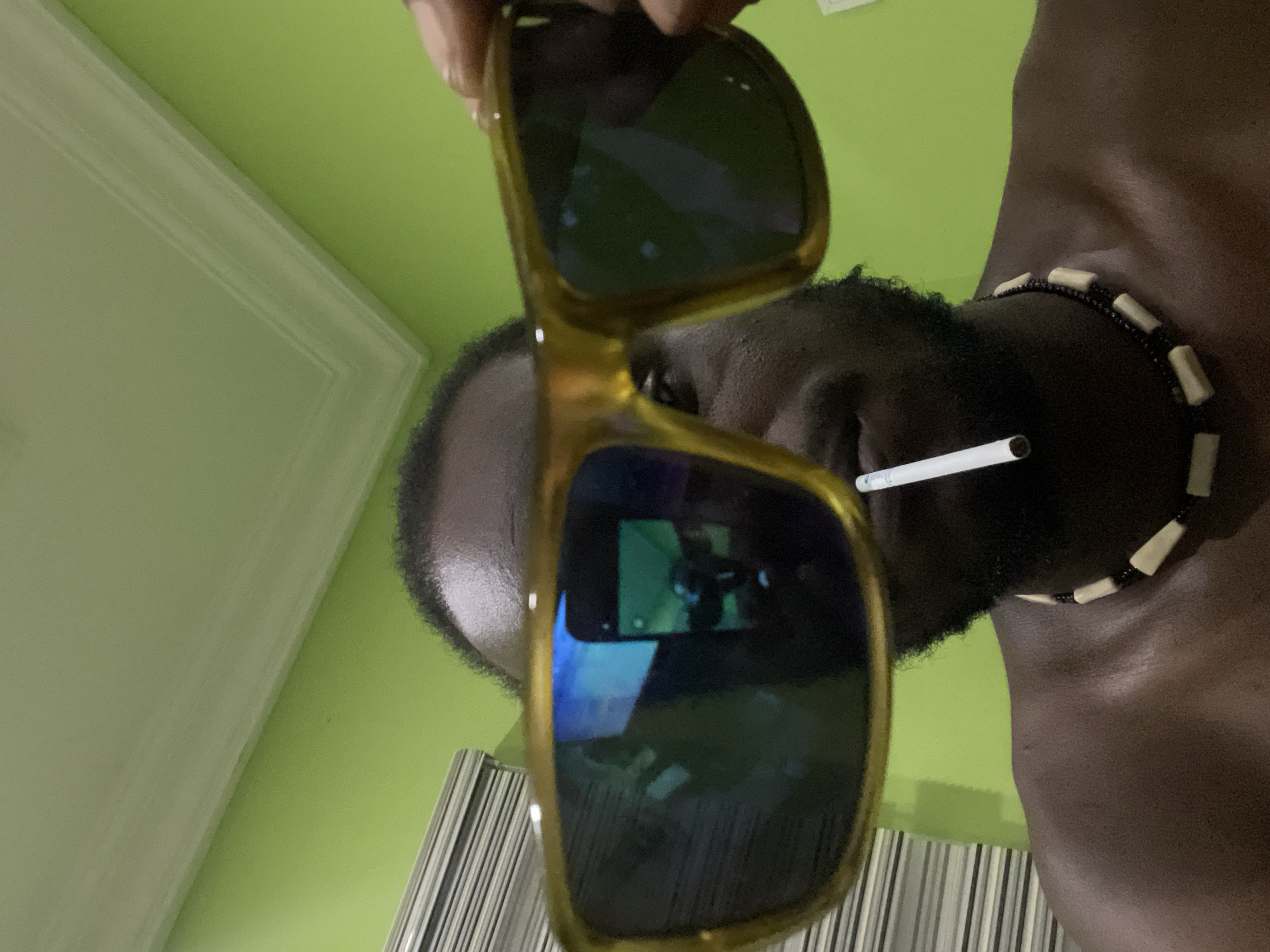 The image shows a person with dark skin and a black beard. A white cigarette is placed in their mouth, and they wear a beaded necklace. The person is holding sunglasses with a yellow frame in front of their face. The background consists of a light green wall and a vertically striped surface. The image does not provide enough detail to confirm the location in Abuja, Nigeria.
