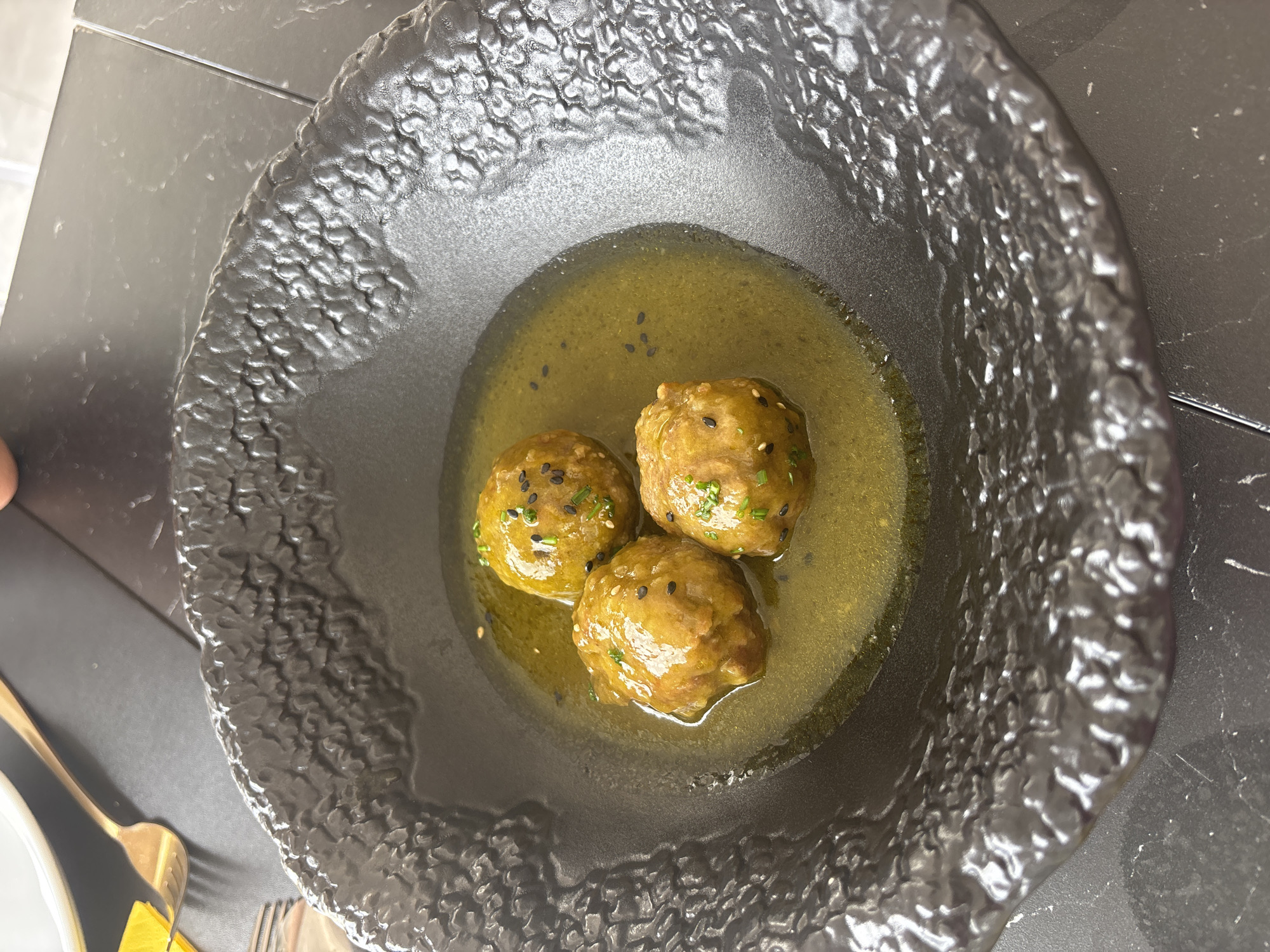 A close-up, overhead shot shows three meatballs submerged in a yellowish-green sauce within a textured black bowl. The meatballs are sprinkled with black sesame seeds and finely chopped green herbs. The bowl is placed on a dark, possibly marble, surface. In the upper left corner, a gold fork and a slice of yellow food, possibly lemon, are visible. The scene appears to be in a restaurant setting at El Parador de las Hortichuelas, Spain. No people are visible.