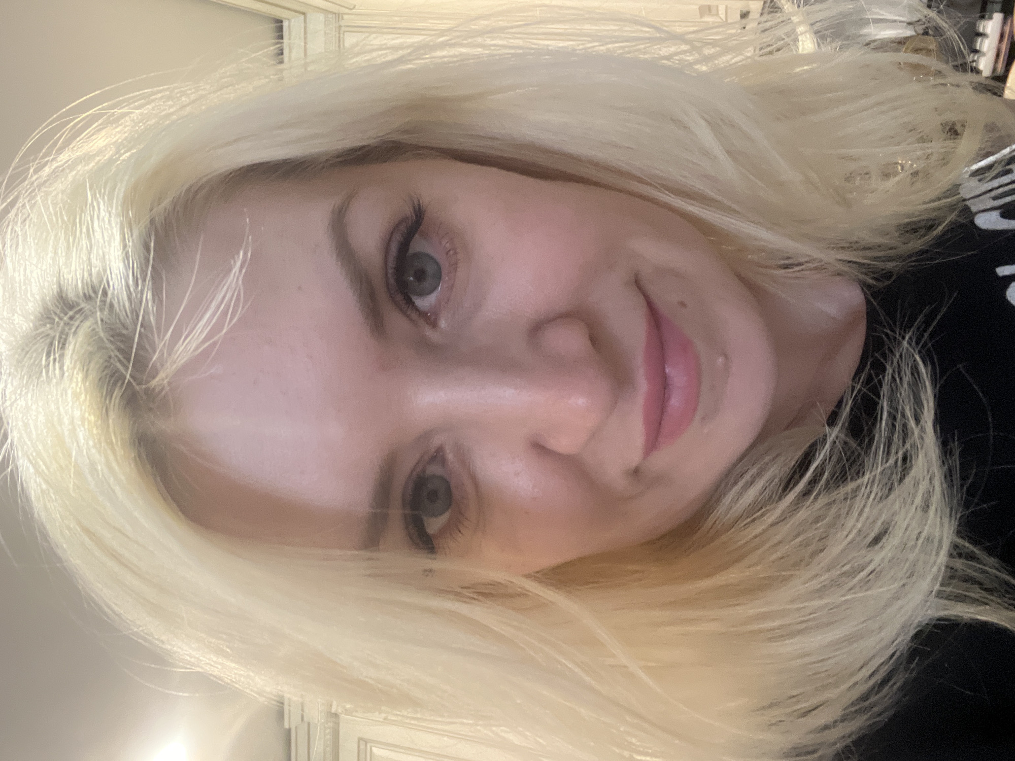 A close-up selfie of a woman with blonde hair and blue eyes is captured. She is wearing a black t-shirt. The background appears to be an indoor setting with what looks like kitchen cabinets and lighting fixtures. The image does not contain any specific landmarks or visual cues that would allow for identification of its location within Tyumen, Russia.