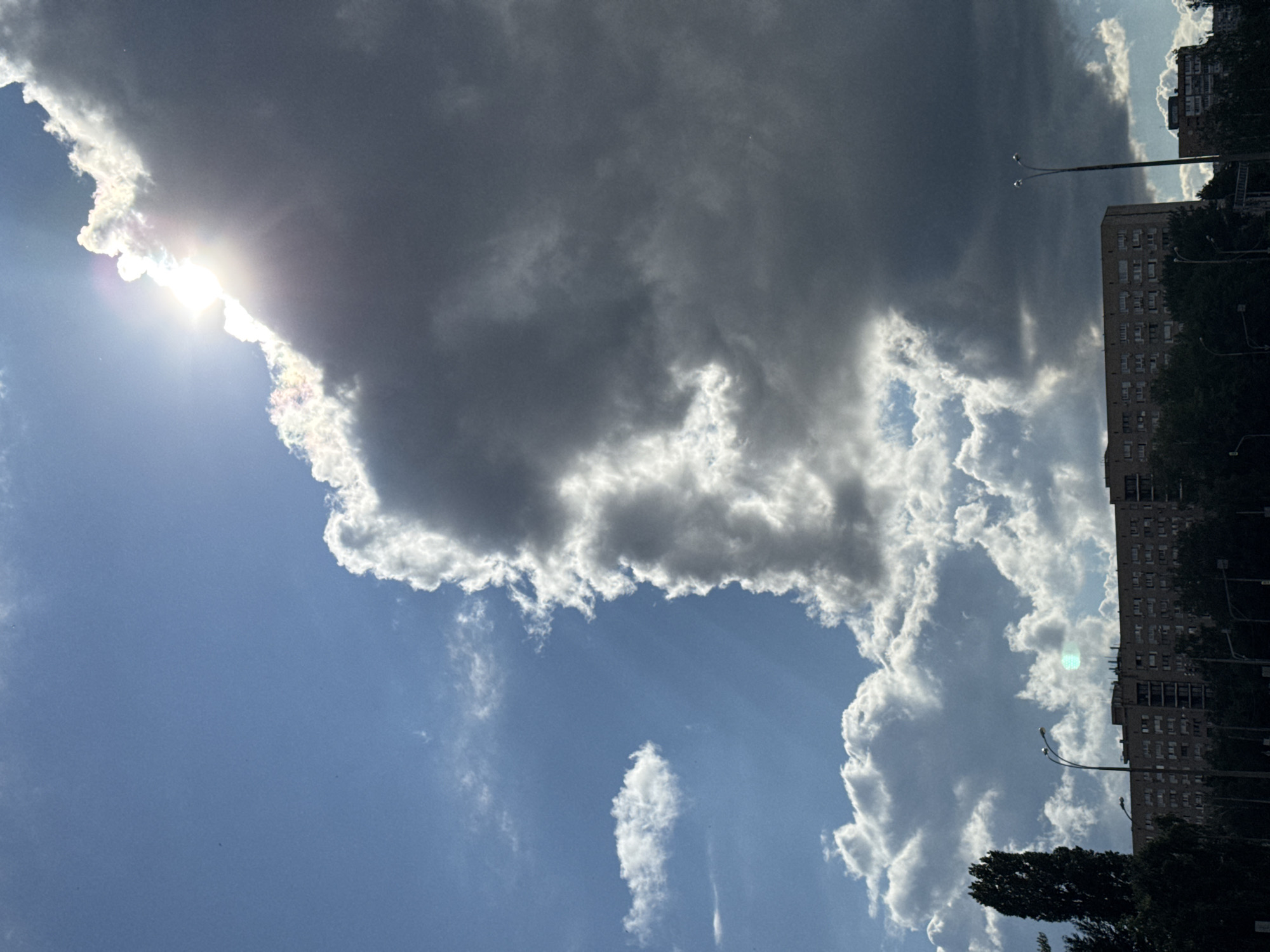 The image depicts a scene in an urban environment, likely a city, characterized by a dramatic sky and a building in the background. The sky is a prominent feature, divided between a bright blue expanse on the left and a large, dark, ominous cloud formation on the right. The sun is partially obscured by this cloud, creating a bright flare and highlighting the edges of the cloud with white light, suggesting it is either rising or setting, or a storm is approaching.
In the foreground and middle ground, a long, multi-story building stretches horizontally across the frame. Its facade is visible, with numerous windows suggesting it is an apartment building or similar structure. Trees are visible in front of and around the building, some appearing as a dense line of foliage, and a tall, slender tree stands out on the left. Streetlights are also present, with at least two visible poles with lamps, indicating the presence of infrastructure for illumination. The overall impression is a slice of urban life under a dynamic sky, where natural elements and man-made structures coexist. The specific location is Unknown, Unknown.