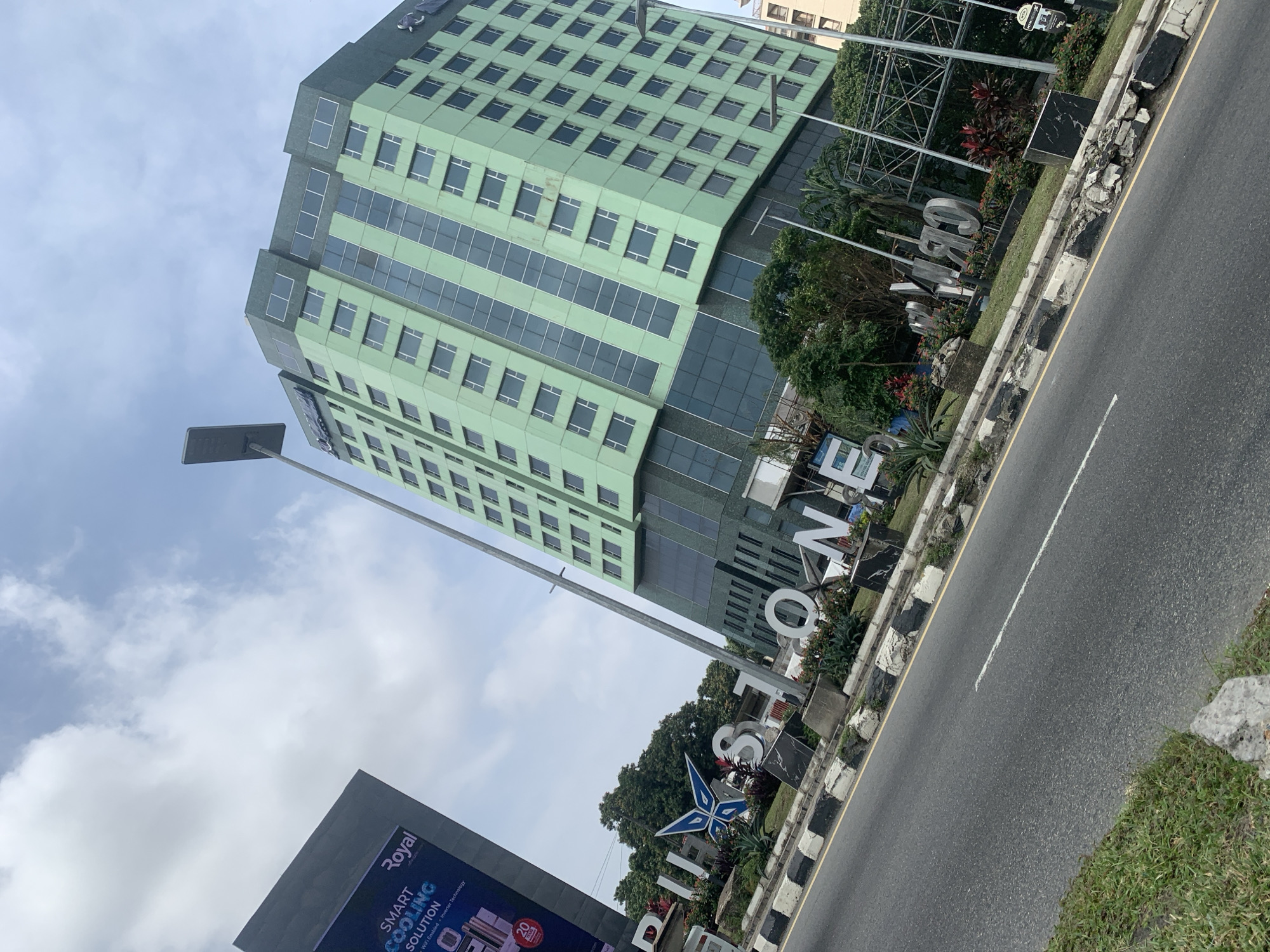 The image shows the "PULSE STONE" signage in front of a tall, light green building with dark tinted windows. The signage is made of large, metallic letters and a blue starburst design. The building appears to be a hotel or office building. To the left of the building, a large digital billboard displays "SMART COOLING SOLUTION" and "Royal Air conditioning". The scene is a street view with a road, curb, and grass on the side. The sky is partly cloudy. The context provided states the location is Ikoyi, Nigeria. There are no people visible in the image.