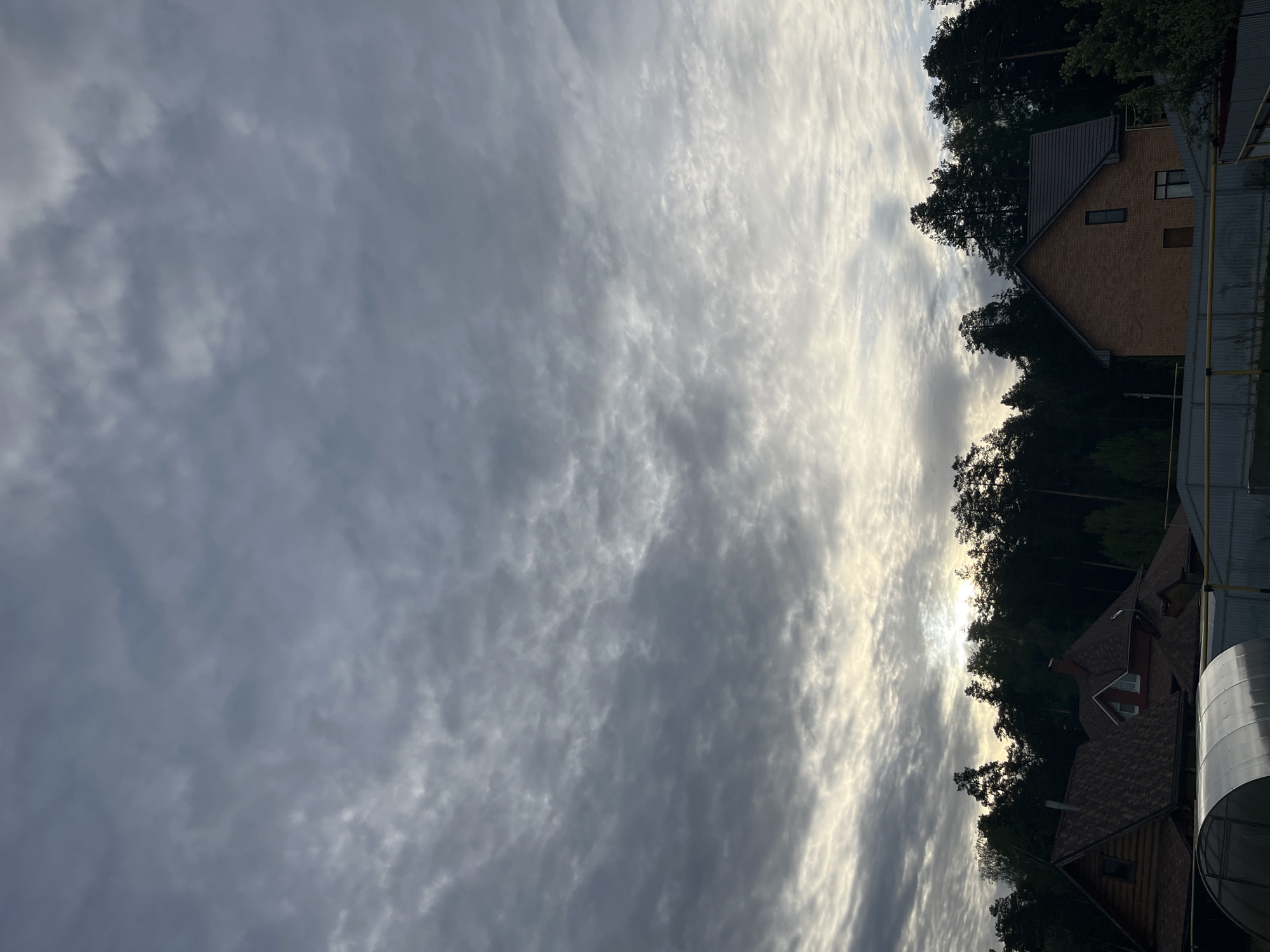The image shows a cloudy sky over a residential area. To the right, houses are visible among trees. The houses appear to be constructed with brick and wood. A metallic cylindrical object is visible on the right side, and there is a metal fence. The image has been rotated, so the sky appears on the left and the ground on the right. It is not possible to confirm the location is in Chaikovsky, Russia, from the image alone.