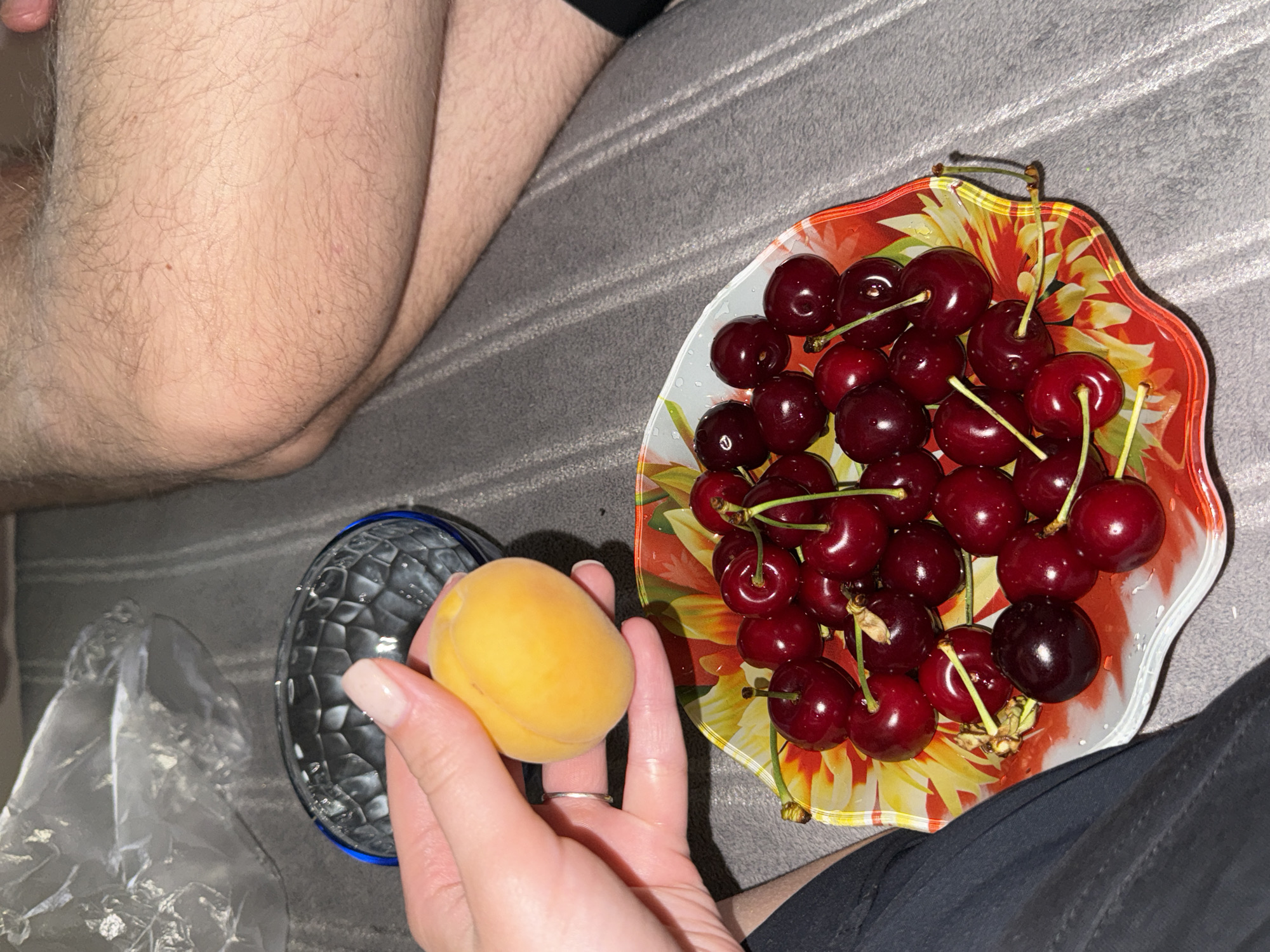 A person's hand holds a single apricot over a colorful, floral-patterned bowl filled with dark red cherries. The hand has light-toned skin, a light pink manicure, and a silver ring on the index finger. The cherries are glossy and appear to be fresh, some still attached to their stems. The bowl is positioned on a gray, textured surface, possibly a blanket or couch. In the upper left corner, a crumpled piece of clear plastic is visible, and to the right, a person's legs, covered in light hair, are seen in the background, suggesting a relaxed, indoor setting. The overall scene is brightly lit, with a shallow depth of field that blurs the background and highlights the fruits.