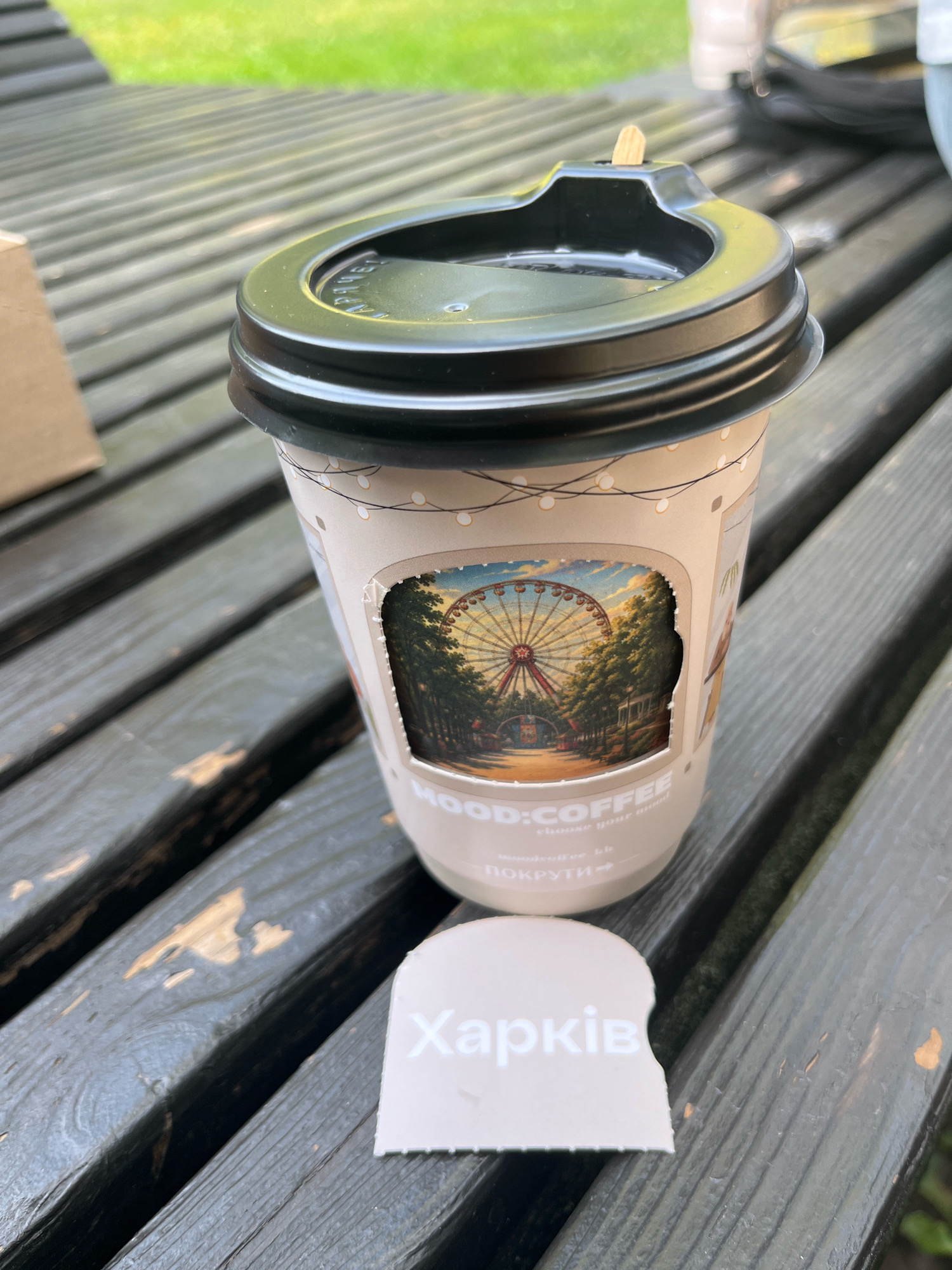 An overhead, close-up shot captures a paper coffee cup with a black lid and a wooden stirrer resting on a dark wooden picnic table outdoors. The cup has a beige sleeve with a cutout revealing a Ferris wheel scene, and text that reads "MOOD:COFFEE" and "choose your mood". A small, torn piece of paper with the word "Харків" (Kharkiv) written in white lettering is placed in front of the cup. The background shows a grassy area.