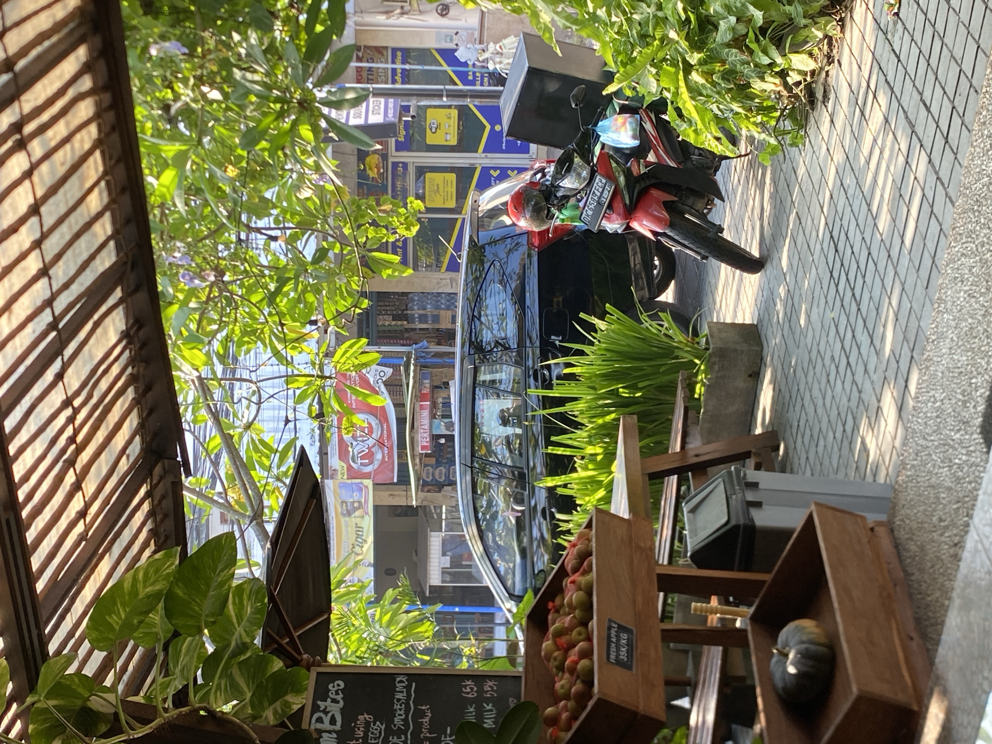 The image captures a street scene in North Kuta, Indonesia, characterized by a blend of commerce and daily life. In the foreground, a wooden display stand holds a crate overflowing with fresh apples, each labeled "FRESH APPLE 35K/KG". Nearby, another wooden box contains a single, small pumpkin. A grey trash bin stands to the right of the display.
The scene is partially shaded by an overhead wooden structure with a translucent canopy, and lush green foliage hangs from above and surrounds the area. In the background, a black car is parked alongside the sidewalk. To its right, a red motorcycle with a large black delivery box on the back is parked, its front tire turned towards the street. A helmet rests on the motorcycle's seat, and a plastic bag hangs from the handlebars.
Behind the parked vehicles, commercial establishments are visible. Signs for "PERTAMINI" (a gas station chain) and "TWIST" (likely a brand) are prominent. The facades of shops display blue and yellow signage with text indicating services like "WI-FI", "PRINTING", "SOUVENIR", and "ADVERTISE". The ground is paved with light-colored square tiles, with a speckled grey textured area in the immediate foreground. The lighting suggests it is daytime, with sunlight casting dappled shadows through the leaves and awning. No people are clearly visible in the image.