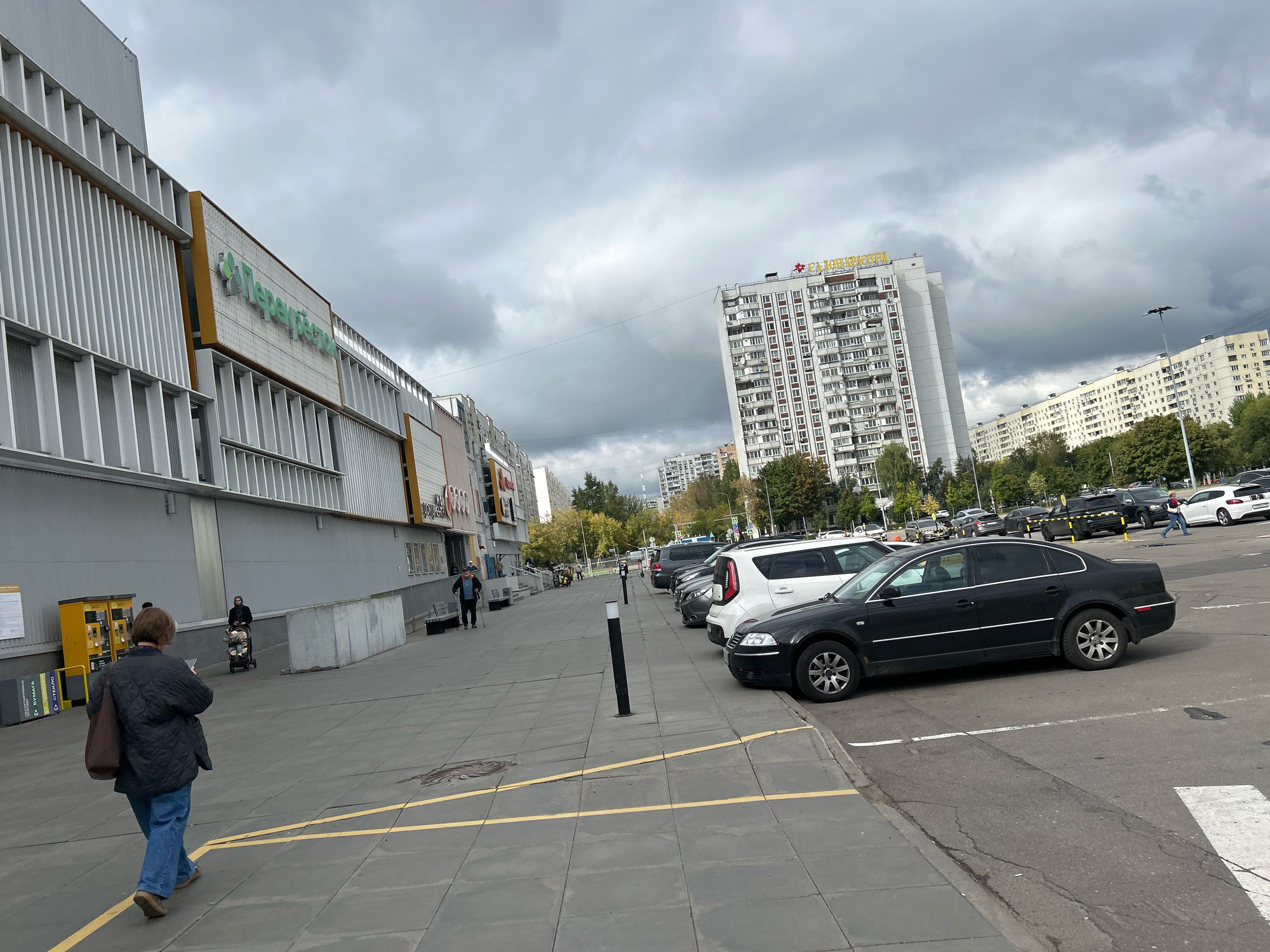 The image shows a parking lot next to a commercial building in Moscow, Russia. The building on the left has a modern facade with vertical louvers and a large sign with green lettering that reads "Перекрёсток", a Russian supermarket chain. In the background, there are several tall, white apartment buildings with many windows, typical of Soviet-era architecture. The sky is overcast with dark clouds, suggesting a possibly gloomy day.
In the parking lot, there are several cars parked, including a black sedan, a white SUV, and other vehicles. People are visible walking on the sidewalk and in the parking lot. One woman in the foreground, wearing a quilted dark jacket and blue jeans, is walking away from the camera. Another person is seen pushing a stroller. An elderly man with a cane is standing near a bench. A yellow parking ticket machine is also visible near the entrance of the building. The ground is paved with gray square tiles, with yellow lines marking parking spaces. The overall scene depicts an everyday urban environment in Moscow.
