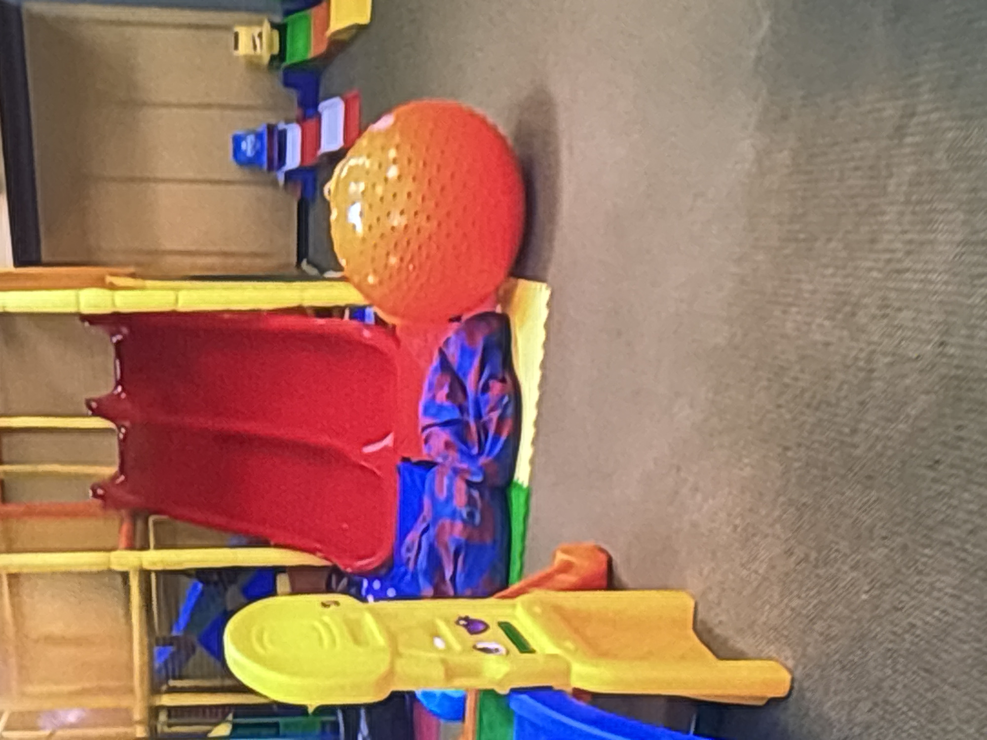 The image depicts a colorful indoor play area. In the foreground, a large, textured orange ball sits on the carpet. To the left, a bright yellow play structure with a smiley face motif is visible, with a blue slide attached to it. Behind these, two large red slides are part of a larger play structure made of yellow and orange plastic. Several beanbag chairs with a blue and purple pattern are scattered on the floor near the slides. In the background, a wall with vertical paneling is visible, with some smaller toy structures or slides, including a blue and white stepped structure and a yellow and green ramp-like structure, placed against it. The overall impression is a child-friendly environment designed for recreation. This scene is located in Dubai, United Arab Emirates.