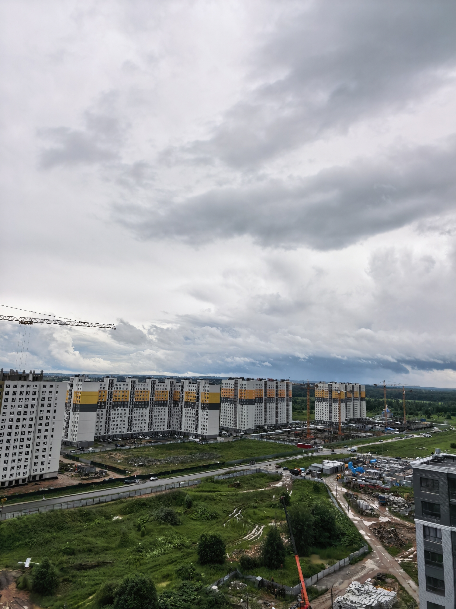 The image depicts a vast construction site in Tver, Russia, under a cloudy sky. In the foreground and midground, several multi-story apartment buildings are under construction or recently completed, characterized by their white facades with yellow and grey accents. Cranes are actively engaged in building operations, with one large tower crane positioned to the left and several others visible throughout the site. The ground is a mix of green, overgrown grass, patches of mud, and areas cleared for construction materials. Piles of building supplies, including what appear to be concrete slabs and construction debris, are scattered across the site. A road runs through the scene, with a few vehicles visible. In the distance, a line of trees forms the horizon, beyond which the landscape appears to be a mix of green fields and more buildings, possibly indicating a developing urban or suburban area. The overall atmosphere is one of ongoing development and industrial activity.