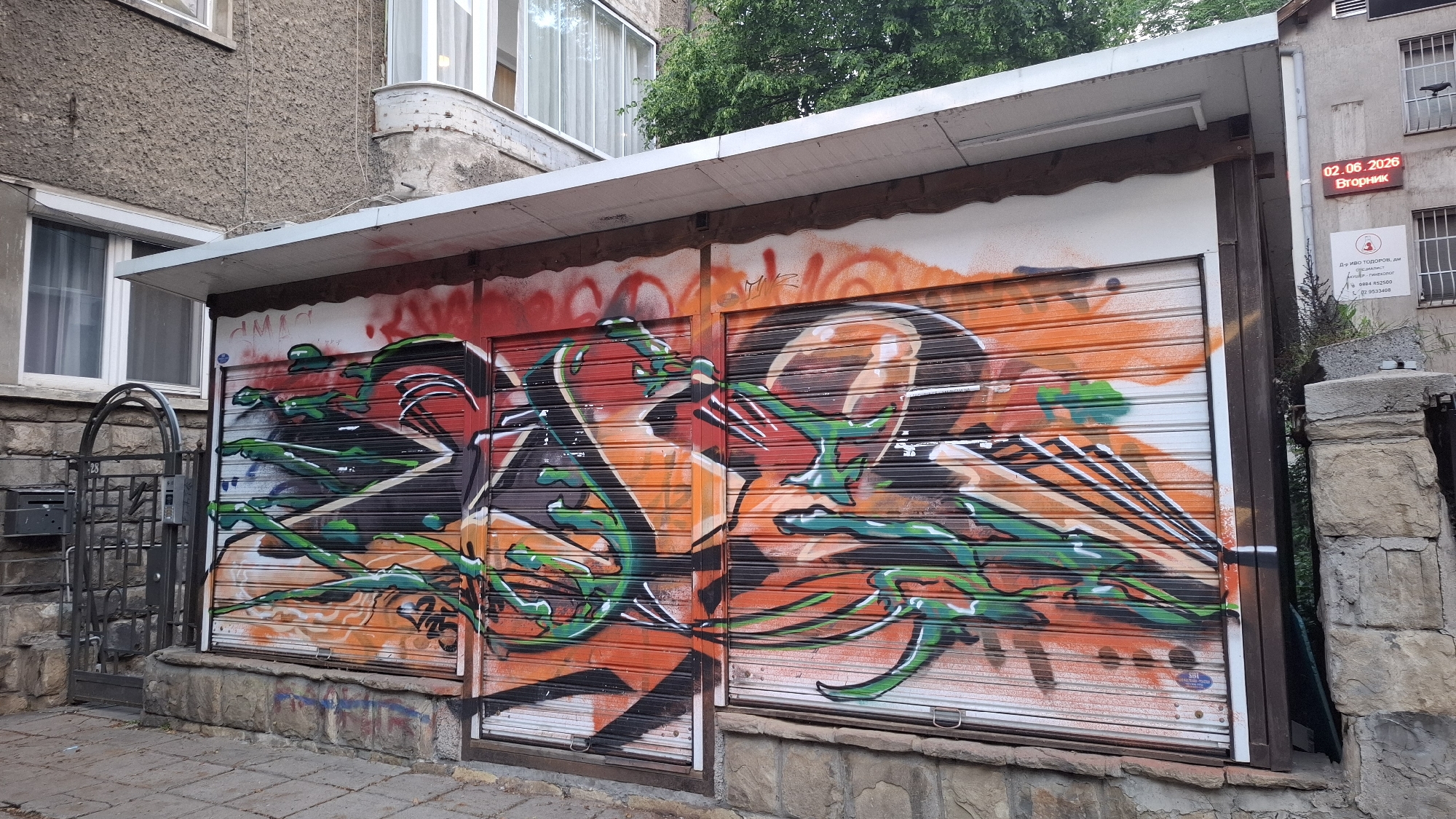 The image displays a vibrant urban scene in Sofia, Bulgaria, characterized by two garage or storage unit roller shutters covered in extensive graffiti. The artwork features bold, abstract designs with flowing lines and organic shapes in bright orange, red, green, black, and white, creating a dynamic contrast against the metal surfaces of the shutters.
Above the graffiti, a white overhang extends from a weathered building with a textured, light gray-brown facade. The building has several white-framed windows; one on the left shows white curtains, while another on the upper right has bars. To the far left, a black metal gate with an ornate arch design and a mailbox is visible, marked with the number "28". The foreground shows a paved sidewalk made of square tiles.
On the right side of the building, a red digital display shows the date "02.06.2026" and the day "Вторник" (Tuesday). Below this, a white sign with black Cyrillic text is visible, reading "Д-р ИВО ТОДОРОВ, дм" (Dr. IVO TODOROV, MD) and a phone number "тел. 02 953408", suggesting a medical practice. A small, dark bird is perched on a windowsill higher up on the building to the right. Green tree foliage is visible in the upper background. The scene is set during daytime, with diffused lighting suggesting an overcast sky or late afternoon.