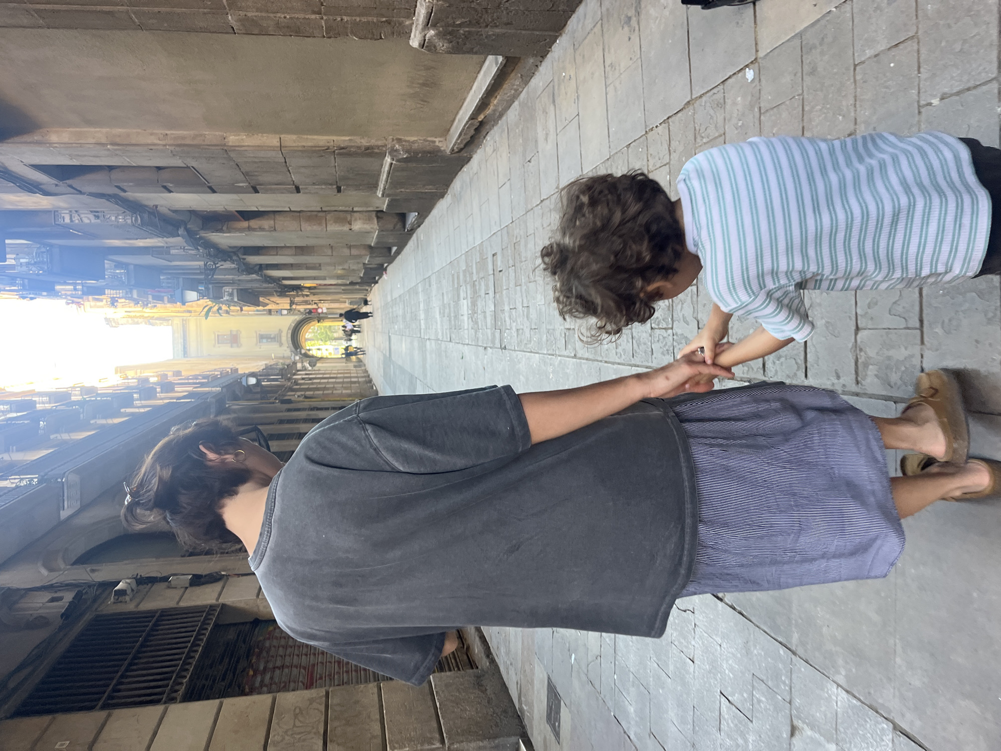 A medium shot shows two people, one adult and one child, holding hands in a narrow stone passageway. The passageway is flanked by buildings with architectural arches, reminiscent of the Gothic Quarter in Barcelona, Spain. The adult, facing away from the camera, is wearing a grey t-shirt and a blue striped skirt. The child, facing the adult, is wearing a white and blue striped shirt and dark pants. The passageway extends into the distance with more people visible in the background, leading to a brighter area. The stone pavement is composed of rectangular blocks. Buildings flank the passageway, with architectural details such as arches and windows.