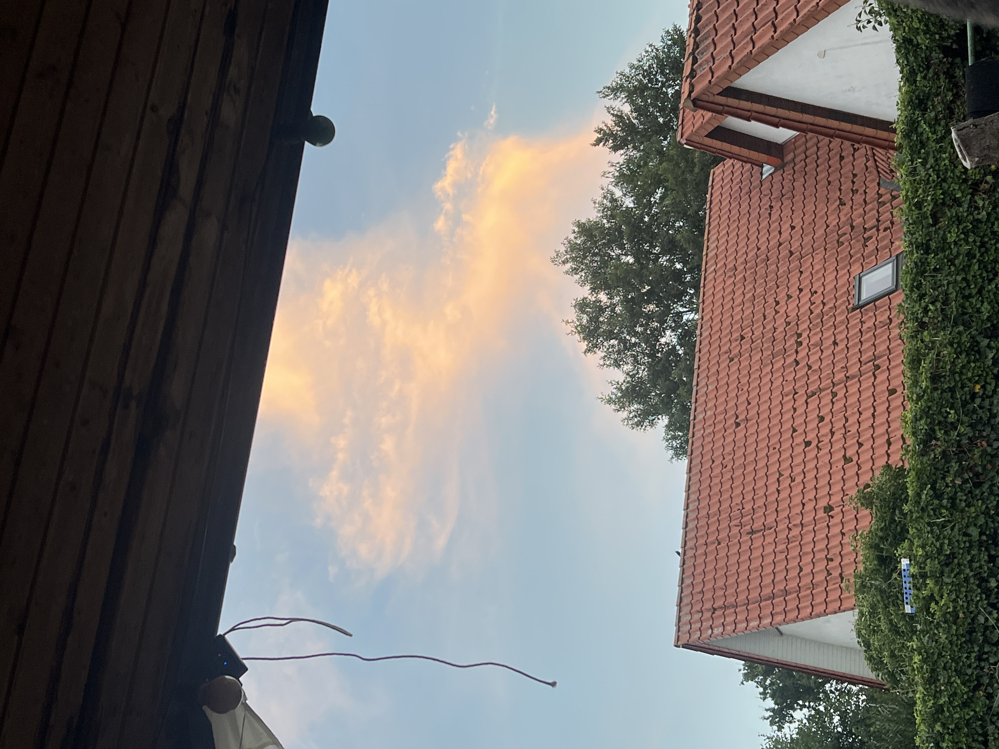 The image captures a view of rooftops and the sky from under a wooden overhang. The sky is partly cloudy, with a large, elongated cloud illuminated by soft, peachy light, suggesting either sunrise or sunset. Below the sky, the red-tiled roofs of houses are visible, partially obscured by lush green hedges. The architecture appears to be of a residential nature, with one house featuring a dormer window and another a skylight. Trees with dark green foliage are seen behind the houses. A thin, dark string hangs down from the overhang on the left side of the frame. On the right side of the overhang, a round object, possibly a light fixture or decoration, is attached. The overall scene evokes a peaceful, domestic setting.