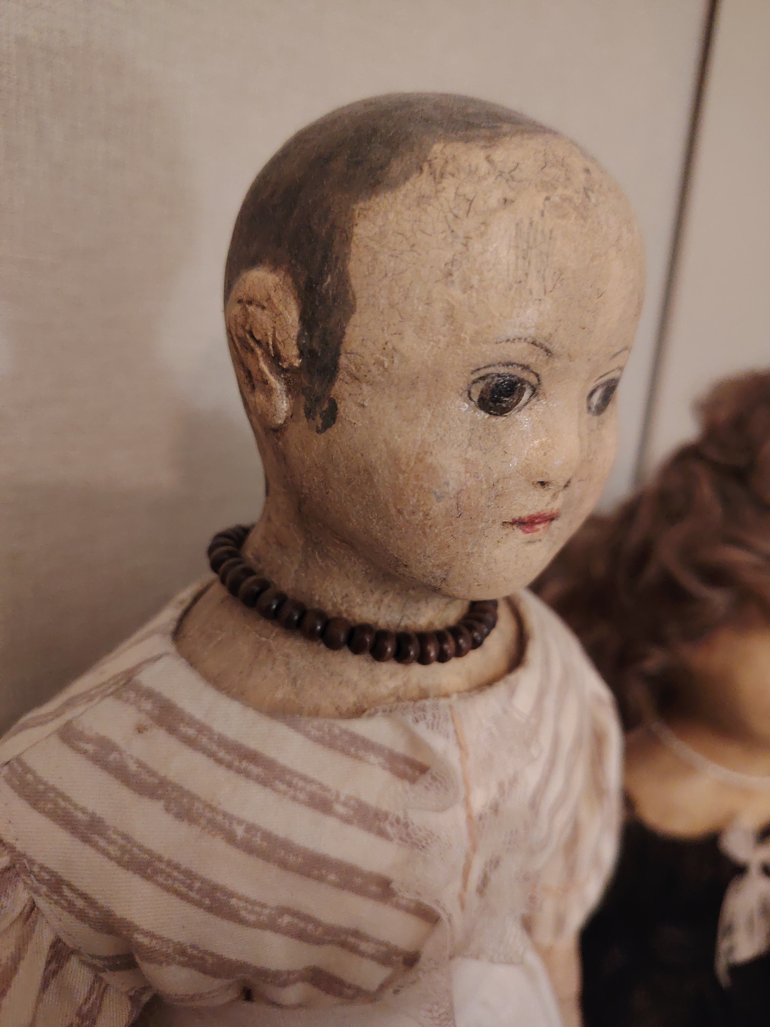 An antique doll is featured in a close-up, slightly off-center portrait. The doll occupies the left and central portion of the frame, with another blurry doll visible in the background to the right.
The primary doll has a head made of a light, aged material, possibly wood or papier-mâché, showing some wear and texture. It features a painted, dark brown cap of hair on the top and sides of its head, with the remainder of the scalp being the natural, lighter color of the head material. Its face has distinct, dark, painted eyes that gaze slightly downwards and to the right, with delicate painted eyebrows. A small, subtly red mouth completes its features. A dark brown, beaded necklace encircles its neck.
The doll is dressed in a garment with wide, horizontal stripes in shades of cream and faded brown on the upper bodice and shoulders. The lower part of its attire, visible towards the bottom right, appears to be a lighter, possibly white or off-white fabric, hinted to be possibly delicate or lacy. The second doll in the background is out of focus, showing only a suggestion of curly, brown hair.
The scene is indoors, against a plain, light-colored background. No specific activity is taking place; the dolls are static objects on display. There is no visible text in the image. The location context is Бараўлянскі сельскі Савет, Belarus.
