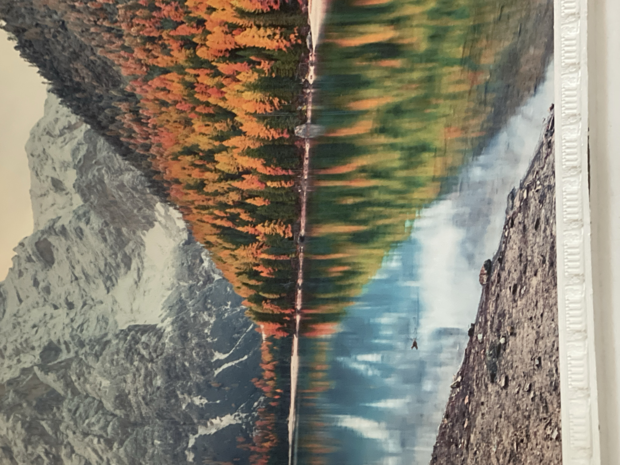 The image depicts a serene mountain lake scene during autumn. A body of water, reflecting the surrounding landscape, occupies the foreground and mid-ground. The lake's surface is calm, with clear reflections of trees displaying vibrant fall foliage in shades of orange, yellow, and green. Beyond the treeline, imposing mountains rise, with their rugged, rocky slopes partially covered in snow. The sky above appears muted, suggesting an overcast day or early morning/late evening light. The shoreline in the foreground is composed of rocky debris and a small patch of sandy beach. There are no people or animals visible in the frame. The location is not specified as being in Bishkek City, Kyrgyzstan.