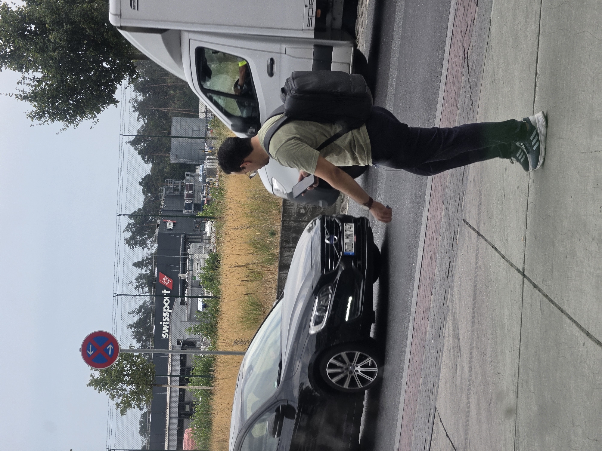 The image shows a street scene in Walldorf, Germany. A man wearing a light green t-shirt, dark pants, and sneakers with a backpack is walking on a sidewalk. He is holding a smartphone in his right hand. A black Volvo car is driving on the road to his left, and a white delivery van is driving on the road to his right. In the background, a building with the "Swissport" logo is visible, along with a traffic sign indicating no stopping in both directions. The sky is overcast, and the weather appears to be mild. The scene suggests a normal day in an urban environment, possibly near an airport or logistics facility given the "Swissport" branding.
