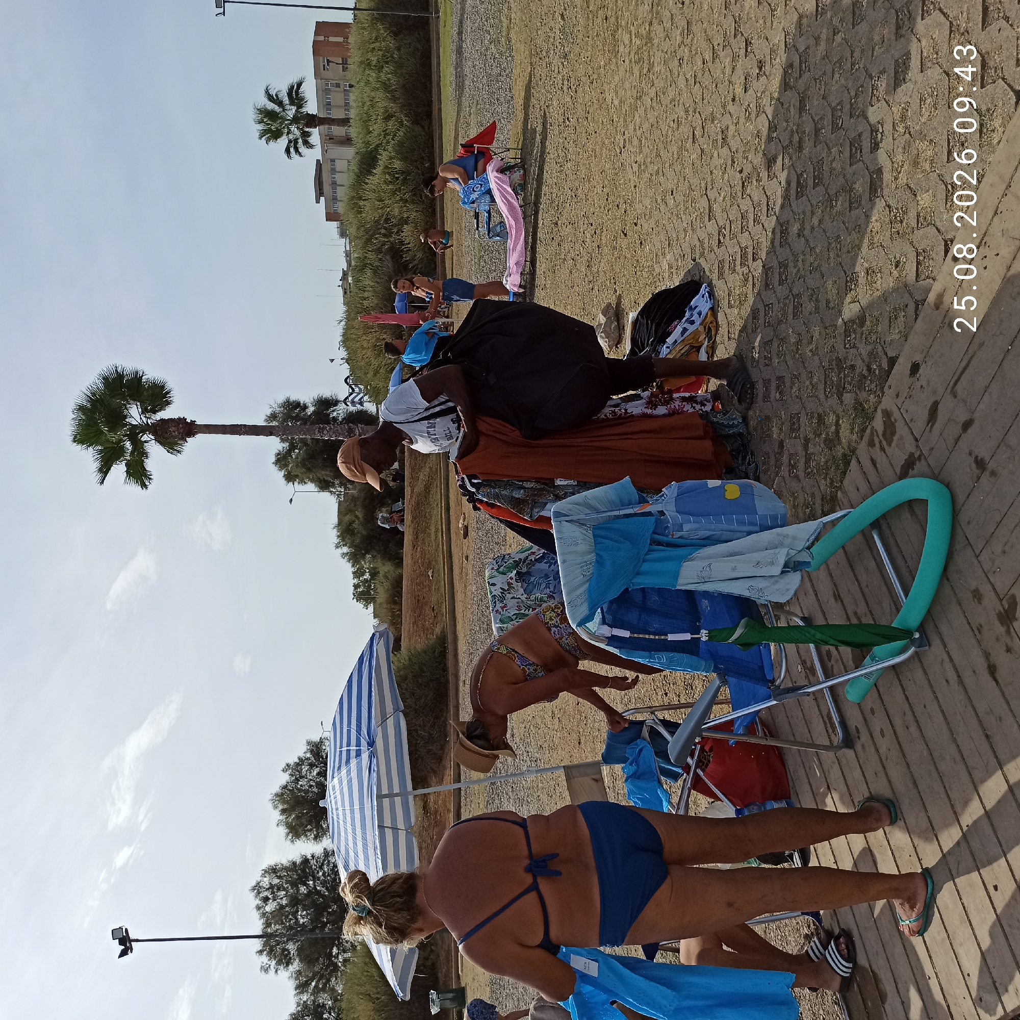 The image depicts a daytime scene at a beach, possibly in Bari, Italy, given the presence of numerous sunbathers and beach paraphernalia. The ground is a mix of sand and interlocking pavers.
In the foreground, a woman in a blue bikini stands on a wooden platform, holding a blue garment. To her right, another woman wearing a straw hat and a patterned bikini bends over near some clothing. Several beach chairs are present, one with a pink towel draped over it, and another holding a blue towel. A blue and white striped umbrella provides shade.
Further back, a man wearing a white t-shirt and an orange cap is seen tending to a rack of clothes, suggesting he might be a vendor. Behind him, a large black bag is visible. Other individuals are scattered across the beach, some sitting on chairs or towels, while others are obscured by vegetation.
The background features dense green bushes and trees, with a palm tree prominently displayed. In the distance, a multi-story building with a light-colored facade and brick accents is visible, along with streetlights. The sky is partly cloudy.
A timestamp in the lower right corner reads "25.08.2026 09:43".