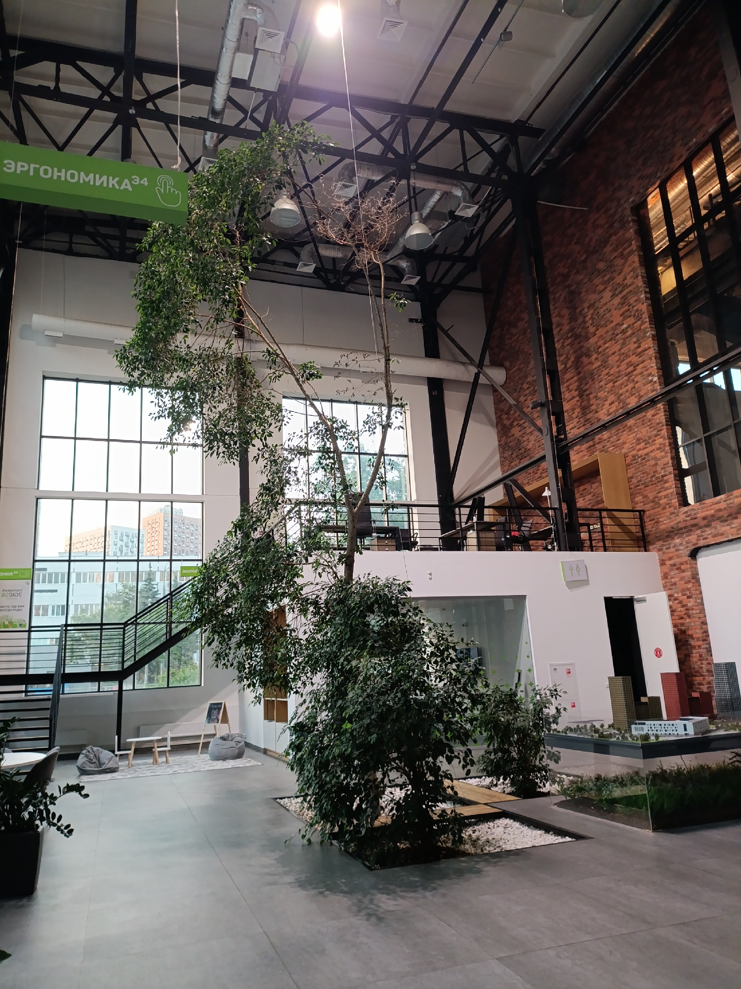 The image shows the interior of a modern, spacious co-working space with an industrial design. A large tree with lush green foliage is prominently featured in the foreground, reaching towards the high ceiling. The space has exposed black metal beams and ductwork, indicative of its industrial aesthetic. Large windows on the left wall offer a view of urban buildings outside. In the middle ground, there is a mezzanine level with offices and seating areas visible. On the ground floor, to the left of the tree, there's a small seating area with beanbag chairs and a low table. To the right of the tree, there's a display of architectural models. The flooring is a light grey tile. A green sign with Cyrillic text, "ЭРГОНОМИКА 34," is visible on the left side, suggesting the co-working space is associated with ergonomics or a company by that name. The overall impression is of a well-lit, contemporary workspace designed for collaboration and comfort.
