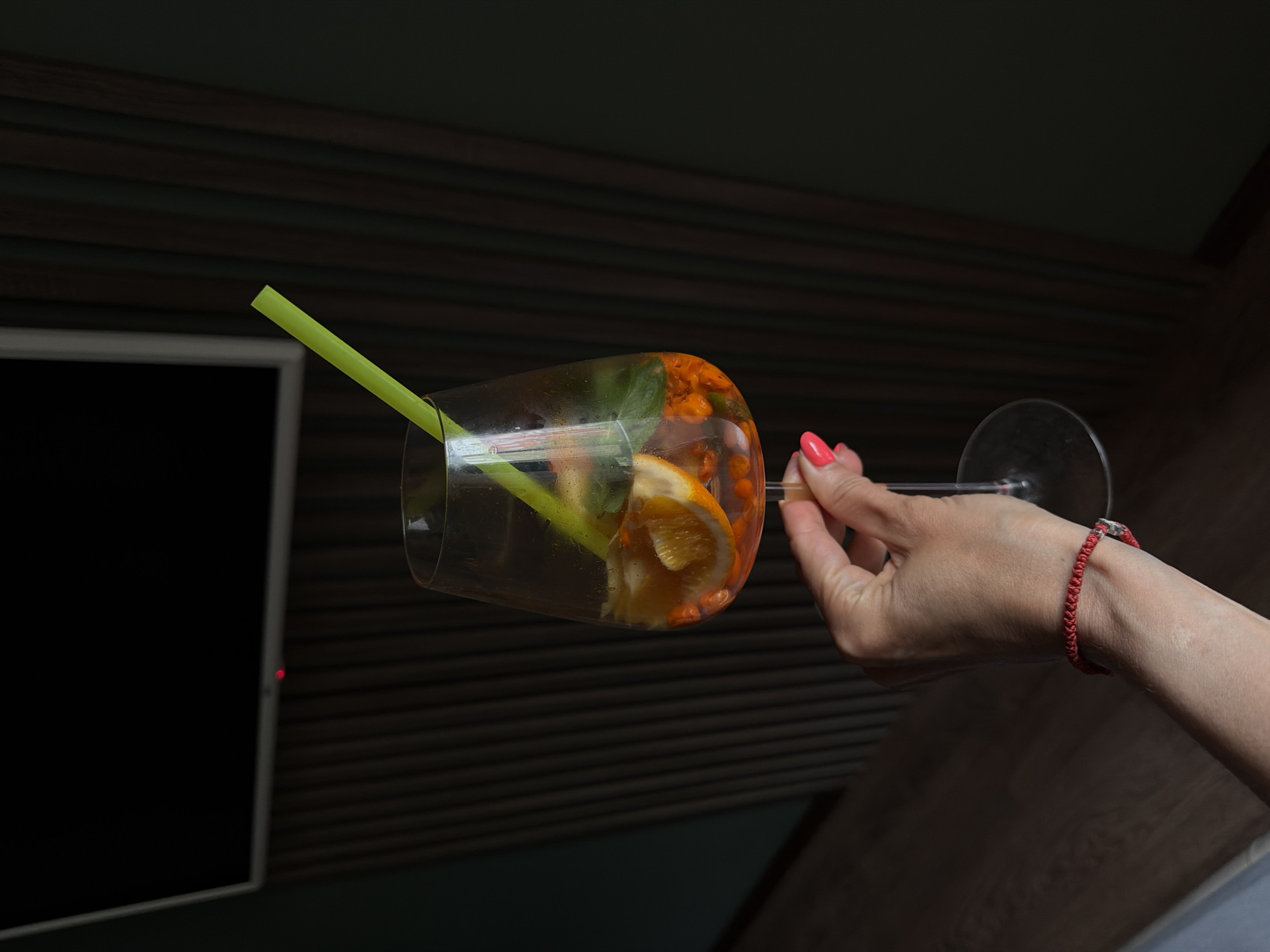 A close-up, vertical shot depicts an individual's right hand holding a stemmed glass indoors, in Birobidzhan, Russia. The hand features short fingernails painted bright coral-pink and a red braided bracelet on the wrist. The glass contains an orange-colored liquid, garnished with sliced oranges, green mint leaves, and small, spherical orange berries, consistent with sea buckthorn. A bright green plastic straw is inserted into the glass. The background consists of a mounted flat-screen television with a dark screen and a visible red power indicator light, situated above a section of dark, vertically slatted or paneled wall. A dark green wall is present to the left, and a very dark wall to the right. A wooden floor with light brown planks is partially visible in the lower left. The environment is dimly lit.