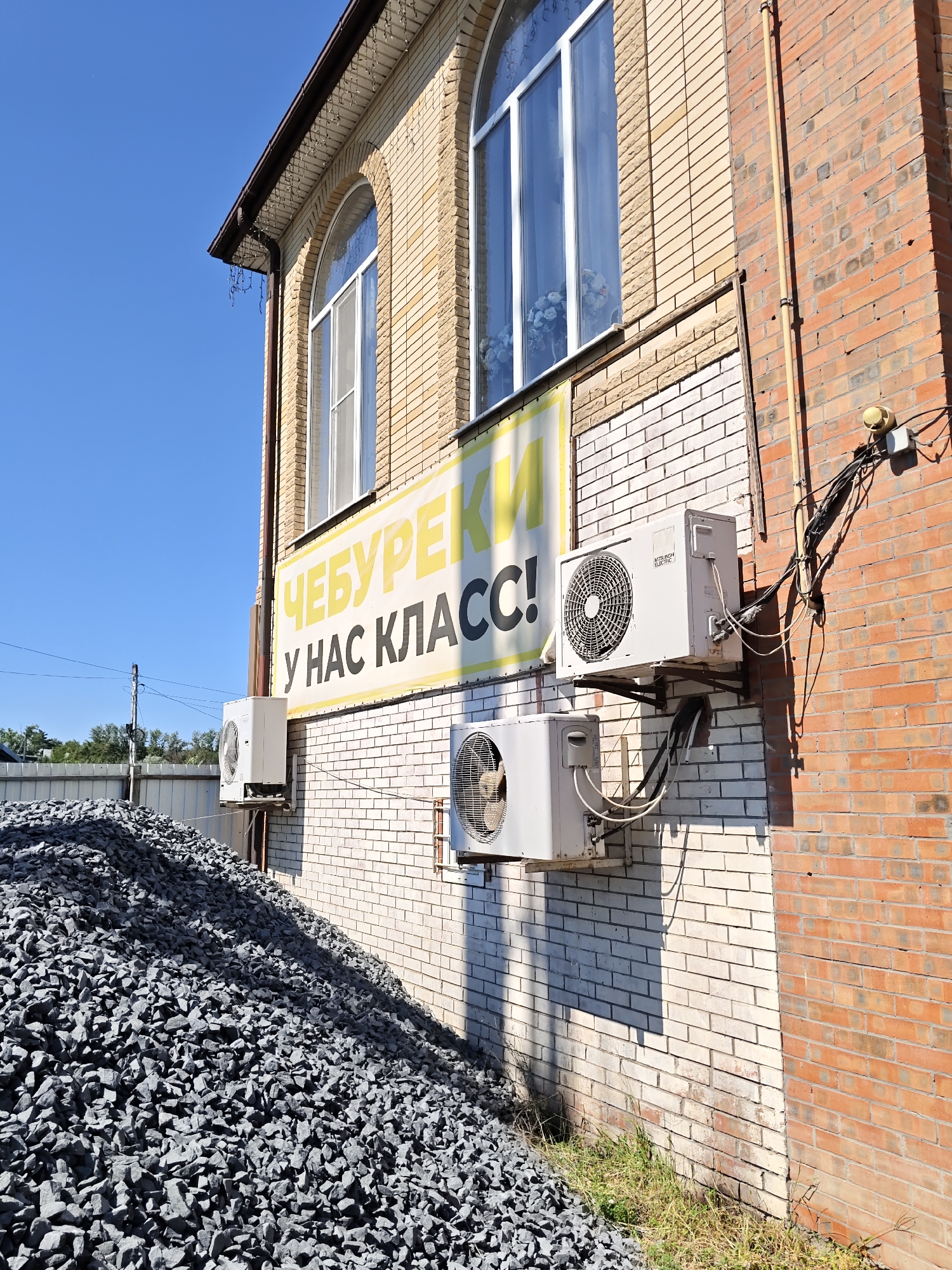The image shows the exterior wall of a two-story building in Rostov, Russia. The building is constructed with a combination of light yellow and red brick. The upper part of the building has arched windows with white frames. A large yellow banner with black lettering is affixed to the wall, with the text "ЧЕБУРЕКИ У НАС КЛАСС!" which translates to "We have class chebureki!". Two outdoor units of air conditioning systems are mounted on the wall beneath the banner. In the foreground, a large pile of dark gray gravel is visible. The sky is clear blue.