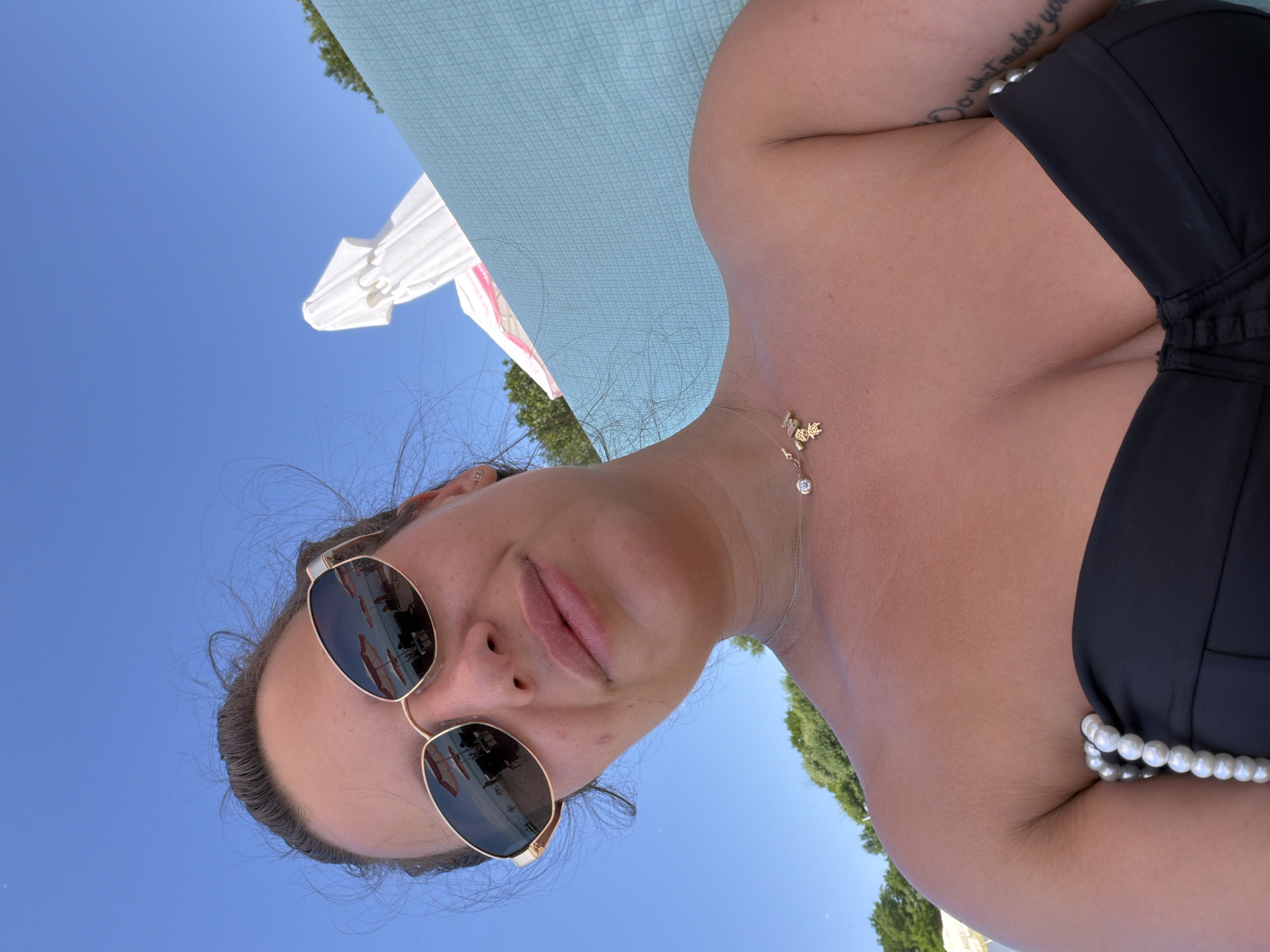 The image shows a woman from the chest up, taking a selfie. She is wearing sunglasses, a black bikini top, and a necklace. The background shows a beach with umbrellas and the sea. The sky is clear and blue. The image is taken from a low angle, looking up at the woman.