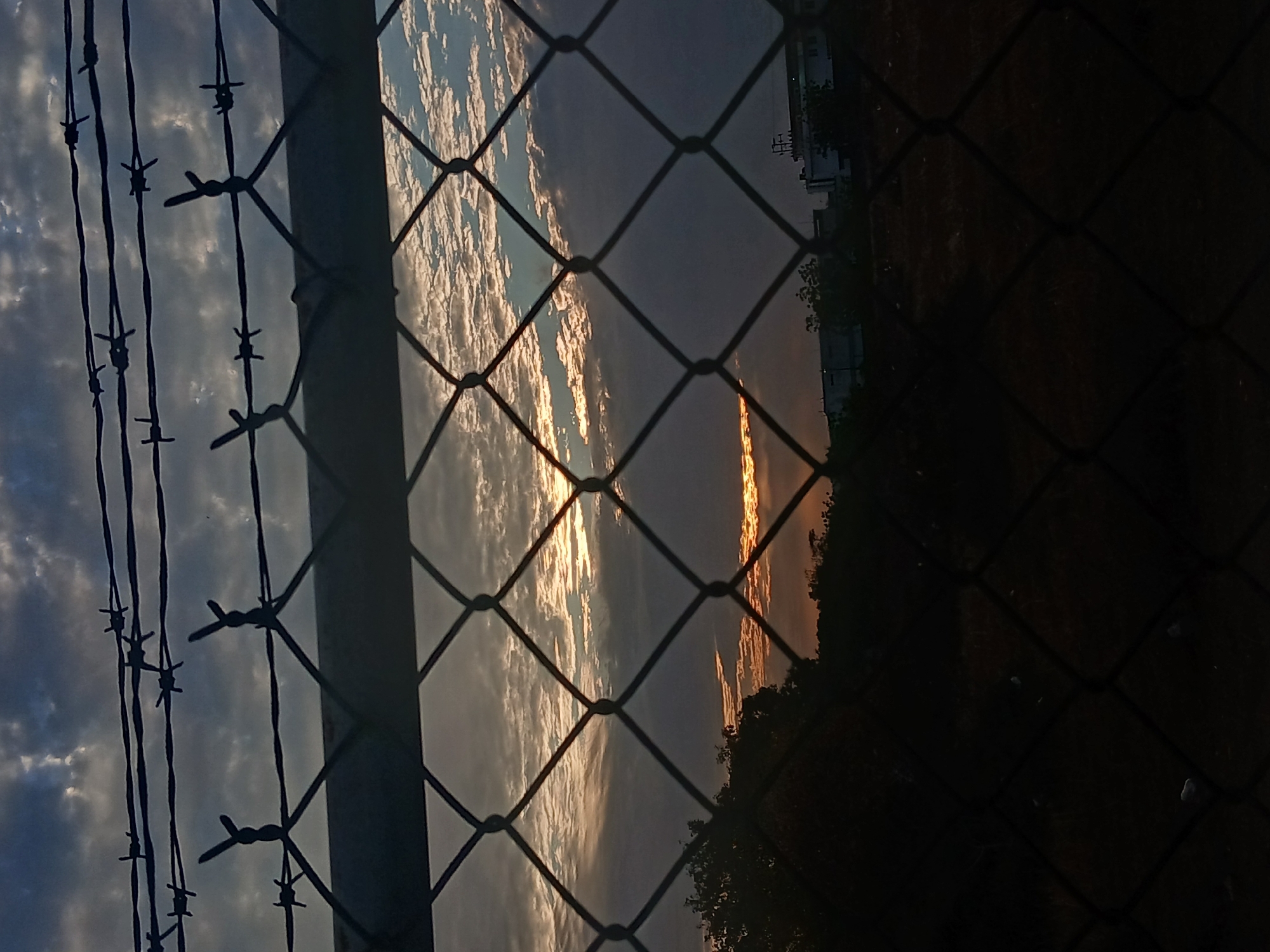 A ground-level, medium, color shot depicts a chain-link fence with barbed wire along the top edge, obscuring the view of the sky and landscape. The photo is oriented vertically. The sky is visible through the chain-link fence, showing a cloudy sky with hints of orange where the sun may be setting. Behind the fence, the land rises and some vegetation can be seen. Buildings are in the distance. The photo was taken in Juan Griego, Venezuela.