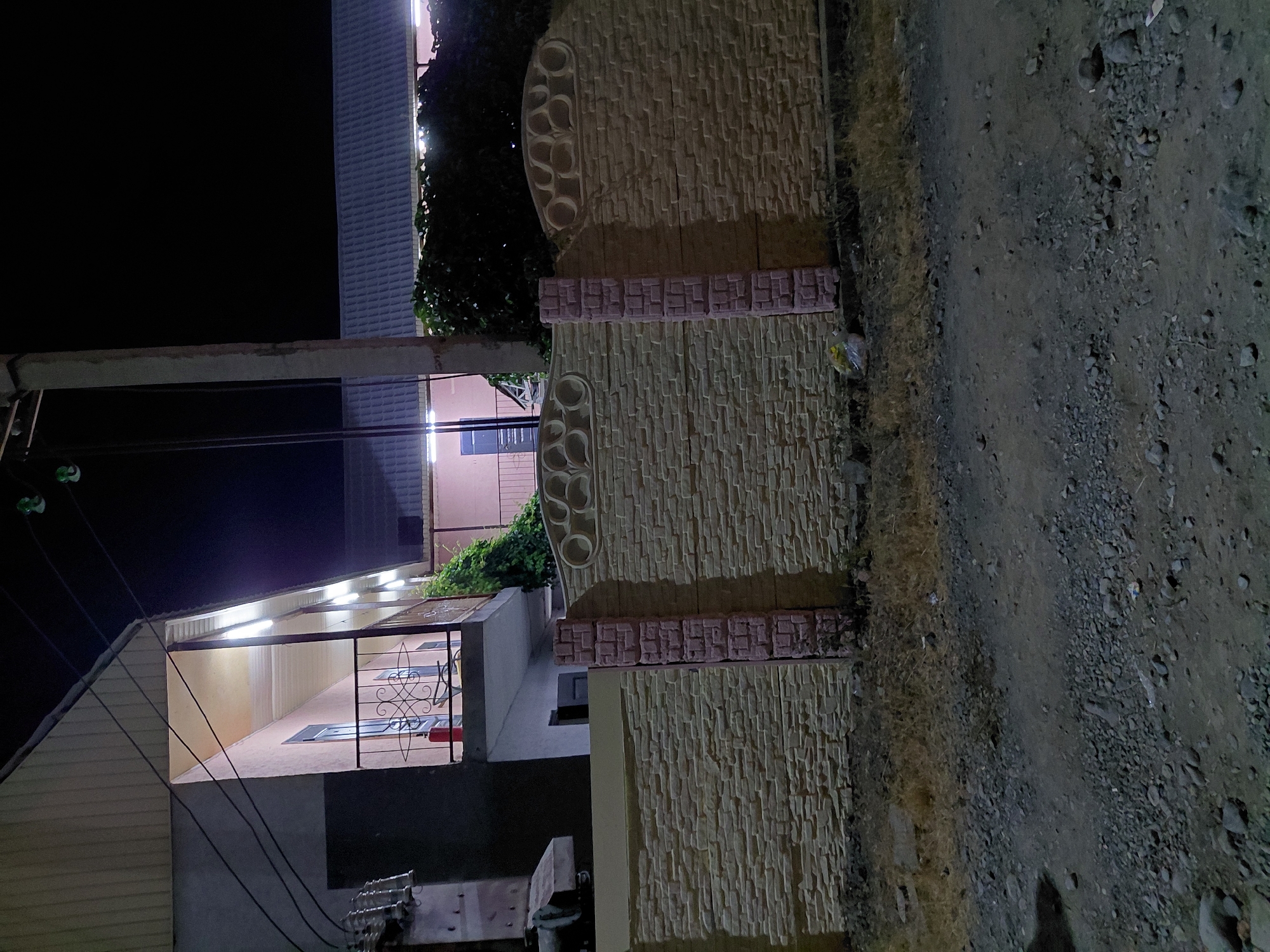 The image shows a nighttime view of a building and a decorative stone fence in Ақши, Kazakhstan. The building appears to be a house or a small business, with a roofline illuminated by bright white strip lights. A covered porch or patio area is visible, with a metal railing and a glimpse of red and blue objects inside. The fence is made of light-colored textured stone with decorative circular patterns and a band of smaller, darker square bricks. To the right of the fence is a rough, gravelly ground surface. The sky is dark, indicating it is nighttime. There are no people visible in the image. The scene appears quiet and still. The overall impression is of a residential or commercial property at night.