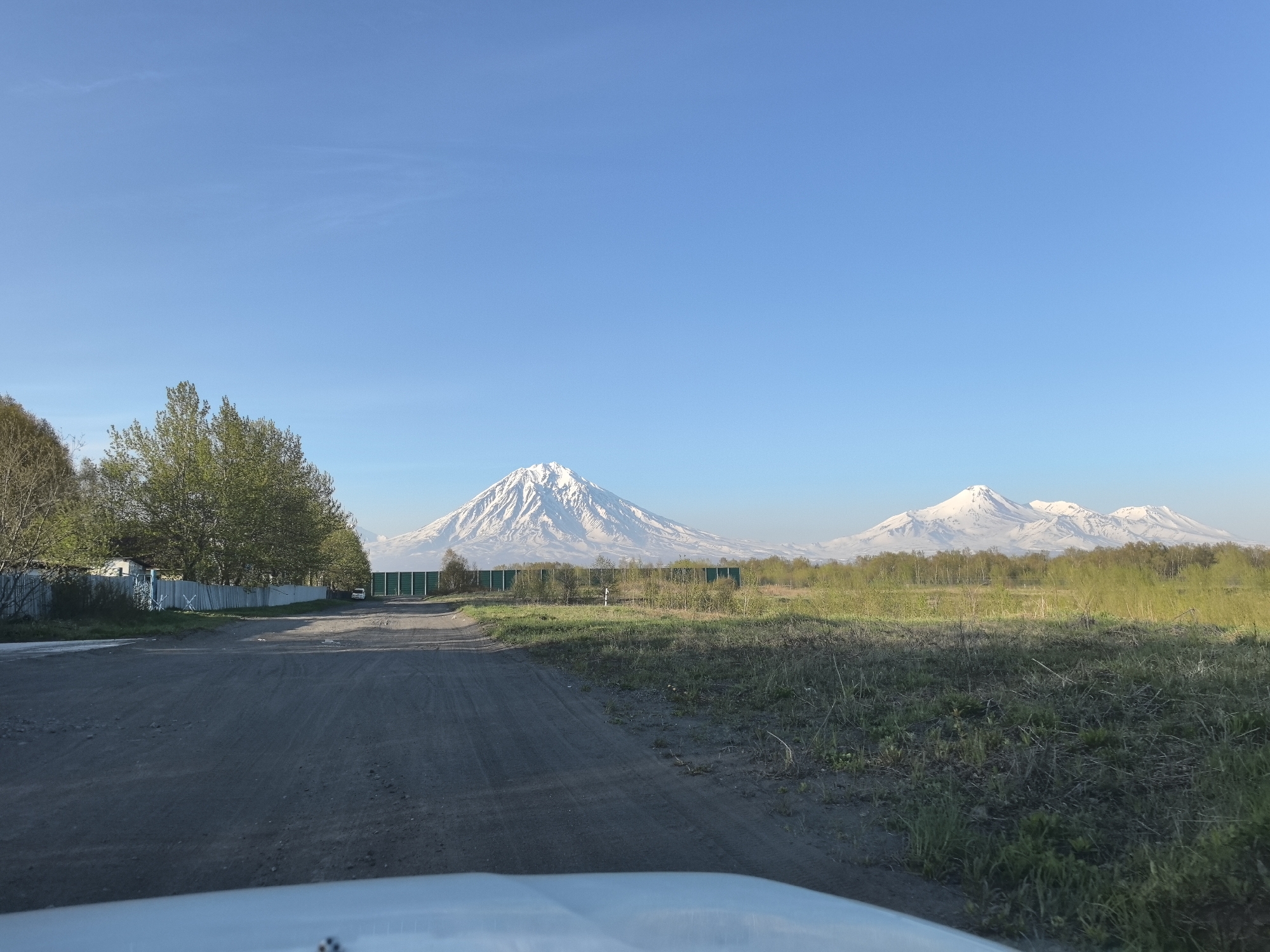 A wide-angle landscape photograph captures a scenic view under a clear, bright blue sky. In the far distance, a prominent chain of snow-capped mountains or volcanoes dominates the horizon. The most striking peak, located centrally on the left, is a tall, symmetrical conical volcano covered in white snow. To its right, several other snow-dusted peaks extend.
The mid-ground features a band of green trees and dense brush. A long, dark green barrier or wall stretches horizontally across the scene, providing a visual separation. To the left, behind a rustic wooden fence, a small white building and part of a white car are visible.
The foreground is dominated by a wide, unpaved dirt road, flanked by patches of green grass and dry brush, curving gently to the right and leading towards the distant mountains. The bottom edge of the frame shows the white hood of a vehicle, indicating the photo was taken from inside a car.
The clear blue sky and bright light suggest a sunny day, likely late spring or early summer, given the vibrant green foliage and the snow still present on the mountain peaks. There is no visible text in the image. The prominent volcanic landscape strongly suggests a region known for active volcanoes, such as the Kamchatka Peninsula. No specific activity is occurring, but the scene implies a journey or a stop along a road.