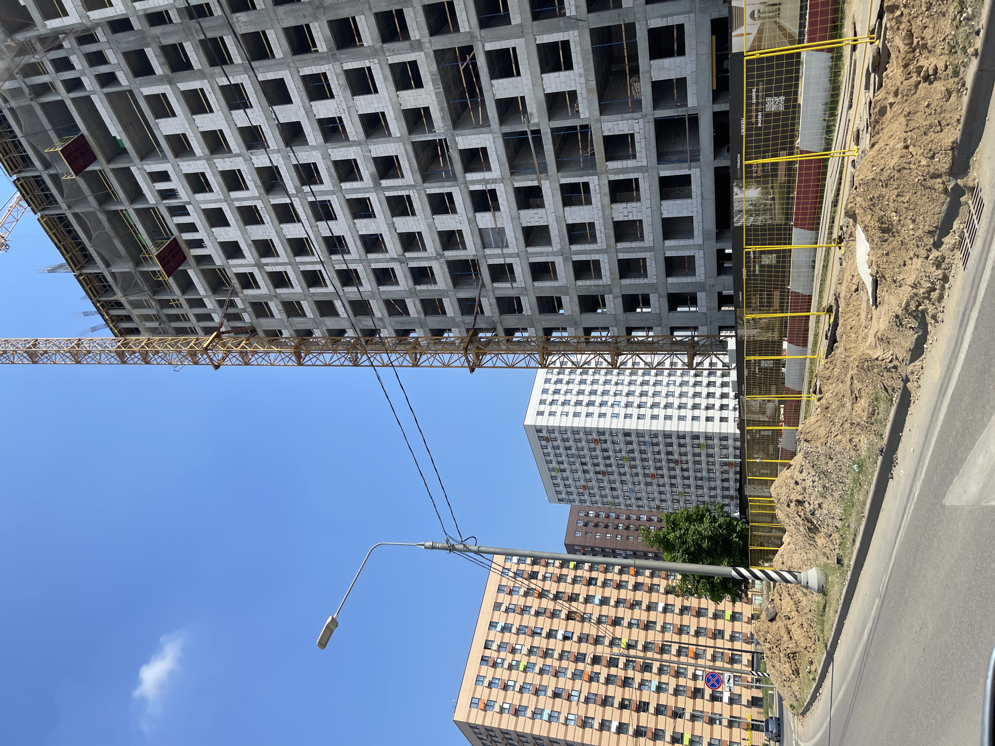 In the city of Kommunarka, Russia, under a clear blue sky with a few wispy clouds, a large construction site is underway. Dominating the right side of the frame is a partially constructed multi-story building, its concrete skeleton revealing rows of empty window frames and balconies. A tall yellow construction crane stands next to it, extending upwards into the sky.
In the mid-ground, two completed residential buildings stand tall. The building on the left is clad in light orange brick with numerous windows, some of which have colorful shutters in shades of green and orange. Adjacent to it is a taller, white building with a grid of windows and colorful accents of green, orange, and red.
The foreground of the image shows a street curving around a construction area. Piles of excavated dirt are heaped along the roadside, separated from the street by a bright yellow safety fence with black banners. A street lamp with a modern design stands to the left of the dirt piles, with power lines running across the scene. A silver car is visible driving on the street in the distance. The overall scene depicts an active urban development environment.