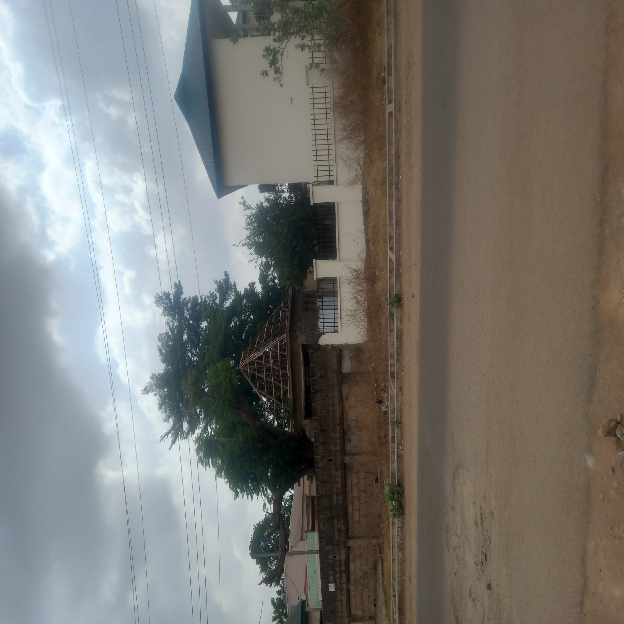 The image captures an outdoor scene along a street in Dadi Riba, Nigeria, under an overcast sky.
The upper portion of the frame is dominated by a cloudy sky, showing patches of brighter light breaking through the grey clouds. Multiple utility wires stretch across the sky from left to right.
On the left side of the street, there is a prominent cream-colored, two-story building with a dark blue or green hip roof. It features several windows, some fitted with dark security bars. In front of this main building, a smaller, unfinished structure, possibly a gazebo or an outbuilding, is visible with an exposed wooden roof frame. Further along the property line, behind a concrete block wall, parts of other residential buildings with light pink and light green facades are partially seen.
Lush, leafy green trees are scattered across the scene, particularly dense around and behind the unfinished structure, contributing significant foliage. Along the edge of the property, separating it from the road, are sparse, dry-looking bushes and weeds.
The right side of the image depicts a wide, unpaved dirt road with a brownish-grey hue. The road surface appears textured and shows some minor indentations. A lighter, narrow strip, possibly a curb or shoulder, runs parallel to the road, defining its edge.
The scene is static, with no people or vehicles visible. The time of day is daytime, under generally cloudy conditions. Faint, illegible graffiti or markings are present on the concrete block wall. The presence of both finished and unfinished structures, along with the dirt road, suggests a developing residential or semi-urban environment.