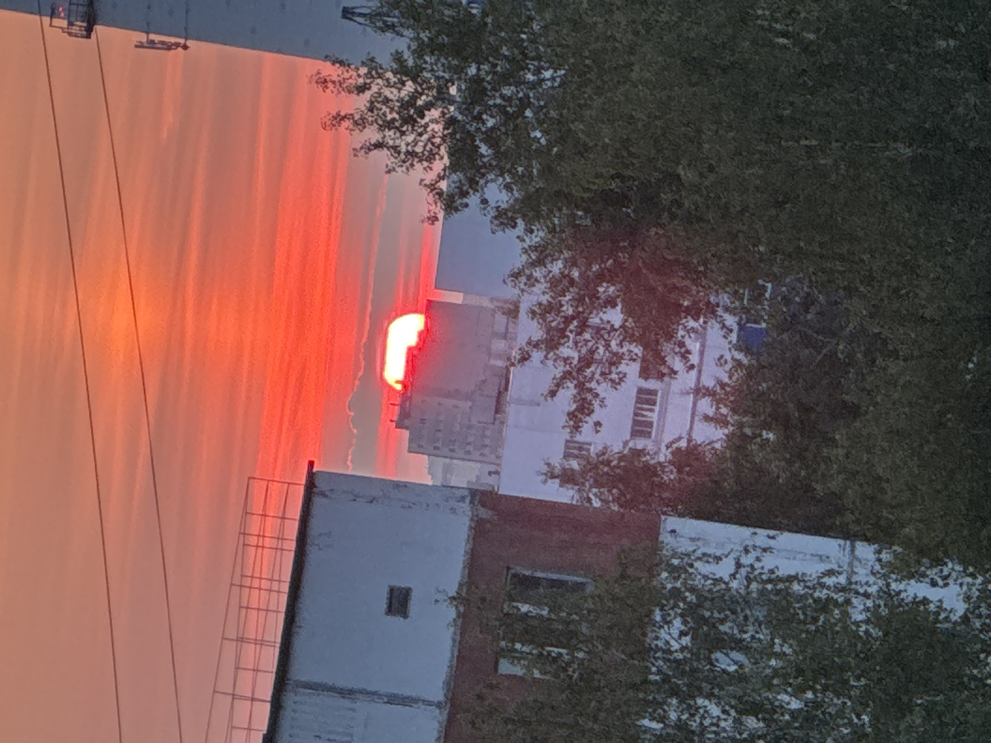 The image captures a sunset over an urban landscape in Surgut, Russia. The sky is dominated by vibrant red and orange hues, indicative of a dramatic sunset. In the foreground, the tops of trees are visible, partially obscuring buildings. Two multi-story buildings are prominent: one on the left with a red-brick lower section and a white upper section, and another white building to its right. The building on the left has several visible windows and a small square opening. Further in the background, several more high-rise buildings are visible, silhouetted against the brightly colored sky. The sun, a bright red orb, is partially obscured by a building in the mid-ground, casting a warm glow over the scene. A tall, white, industrial-looking structure, possibly a power plant smokestack or a radio tower, is visible on the right side of the image. Power lines are stretched across the sky. The overall scene depicts a typical evening in an industrial city, with the natural beauty of the sunset juxtaposed against the urban environment.