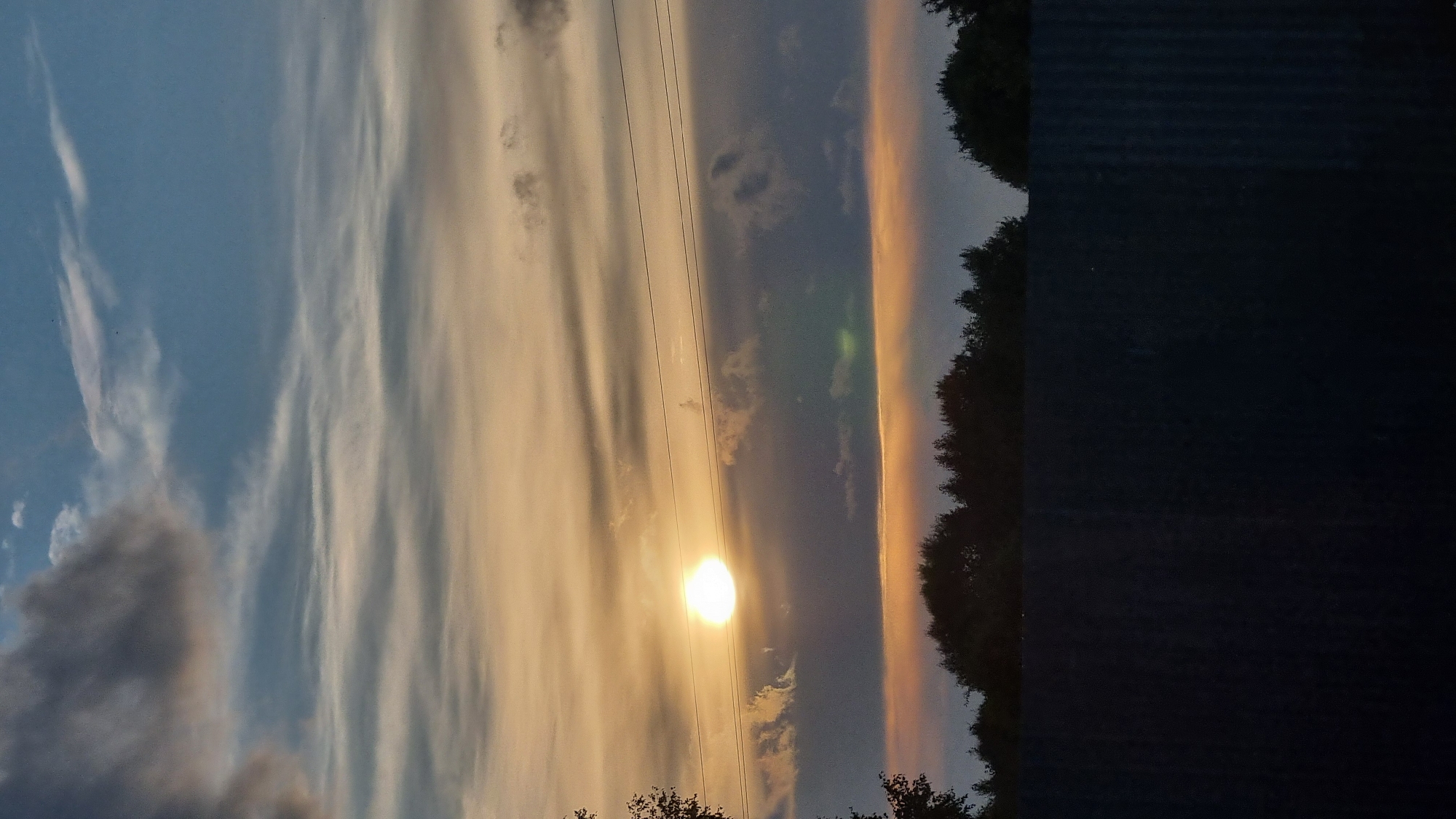 The image depicts a sunset scene over the city of Vysokoe, Russia. The sky is filled with a gradient of colors, from a pale yellow near the horizon to a deep blue at the top. Wispy clouds are scattered across the sky, with some darker, more ominous clouds present. The sun is a bright, glowing orb, partially obscured by clouds. Two power lines stretch horizontally across the frame, bisecting the sky. Below the sky, a dense line of dark trees forms a silhouette against the fading light. In the foreground, a dark, textured fence or wall runs across the bottom of the image. The overall atmosphere is serene and tranquil, with a hint of drama from the stormy clouds.