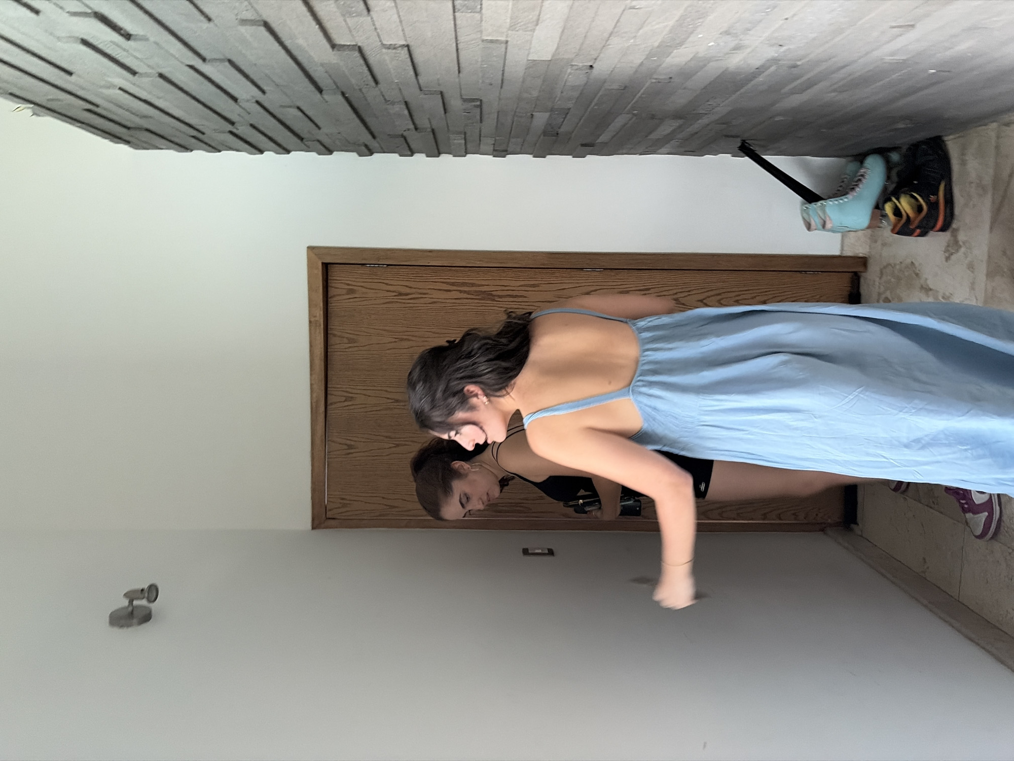 The image, oriented correctly, depicts an indoor scene, likely a hallway or entryway, with two young women. The primary subject, partially seen from the back, is wearing a light blue spaghetti strap dress and has long dark hair. She is leaning forward, looking down, and her reflection is prominently displayed on the highly polished, light-colored floor. The reflection shows another young woman, possibly the same person or a friend, with dark hair tied back, wearing a black top, also looking downwards.
The setting includes a brown wooden door and doorframe on the right side of the image. The wall on the left features distinct gray, rectangular, stacked stone or wood paneling. The floor is a very reflective, light gray or beige surface.
Various objects are visible: a pair of light blue roller skates and a pair of black and orange athletic shoes are resting against the textured wall near the wooden door. A small, silver, cylindrical wall-mounted light fixture is attached to the plain white wall on the left. A purple and white sneaker is partially visible on the floor beneath the blue dress.
The lighting appears bright and even, suggesting an interior during the day. No text is visible in the image. The location is San Bernardino Tlaxcalancingo, Mexico.