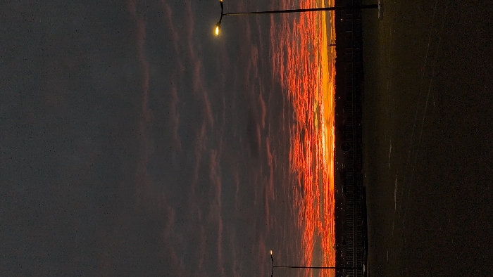 A vibrant sunset paints the sky with fiery hues of orange and red. These colors stretch across a cloudy horizon, contrasted by a dim, gray sky on the left. A slender lamppost stands tall, illuminating a path or field to its right. The ground appears dark, possibly grass or a paved surface, and fades into shadow. The image is vertical and emphasizes the sky's colorful display.
