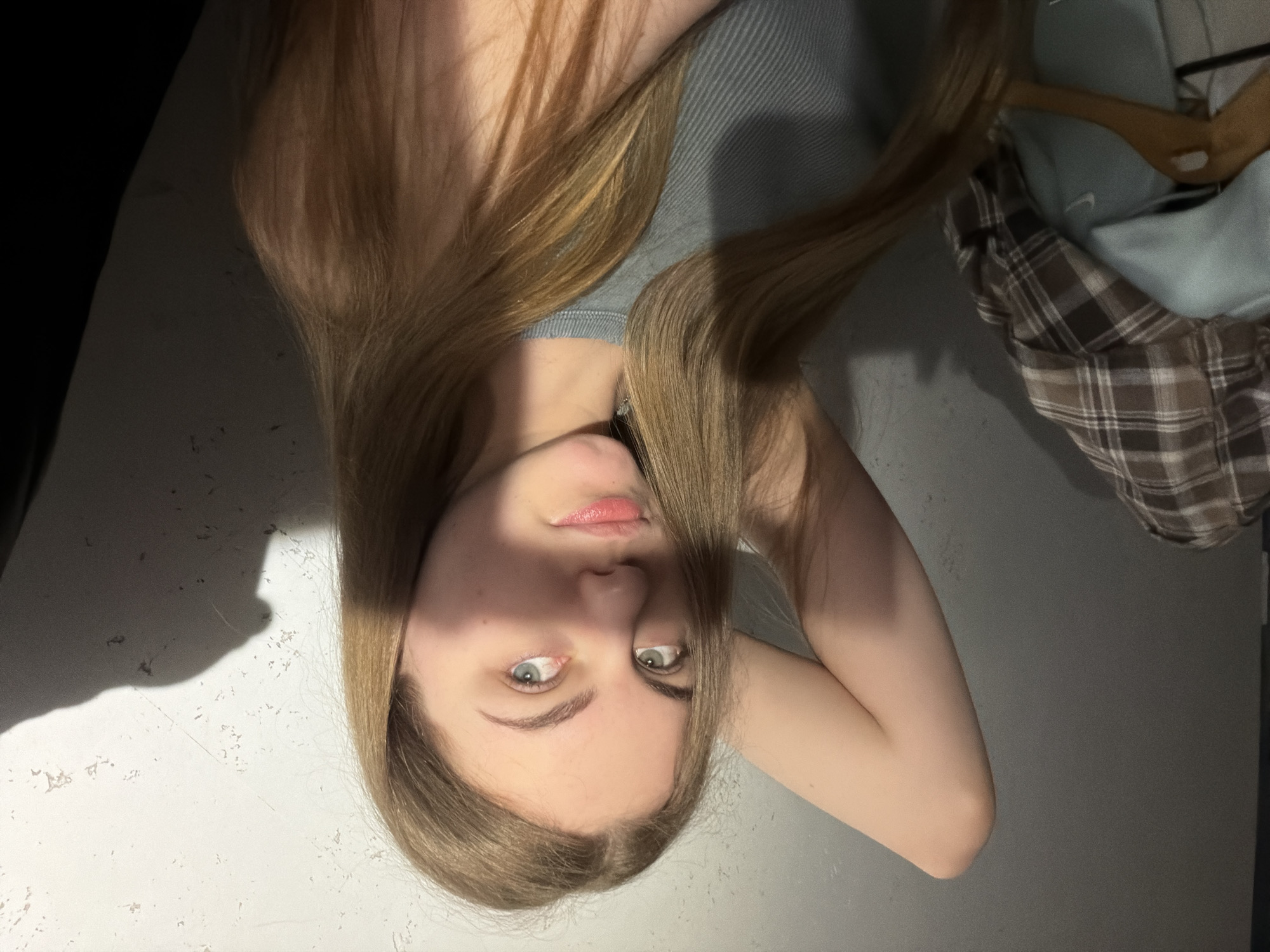 A young woman with long, light brown hair is posing with her left arm raised behind her head. She is wearing a ribbed, grey tank top. Her face is partially illuminated by a beam of light, casting shadows across her forehead and cheek. Her eyes are light blue-green, and her lips are slightly parted. In the background, a grey, textured wall is visible, with a shadow of an object or person cast on the right side. To the left, a stack of folded clothing, including a brown and white plaid shirt and a light blue hooded garment, are arranged on a rack. The setting appears to be indoors, likely a bedroom or dressing area, given the clothing and wall texture. The scene is dimly lit, with the strong directional light creating a dramatic effect.