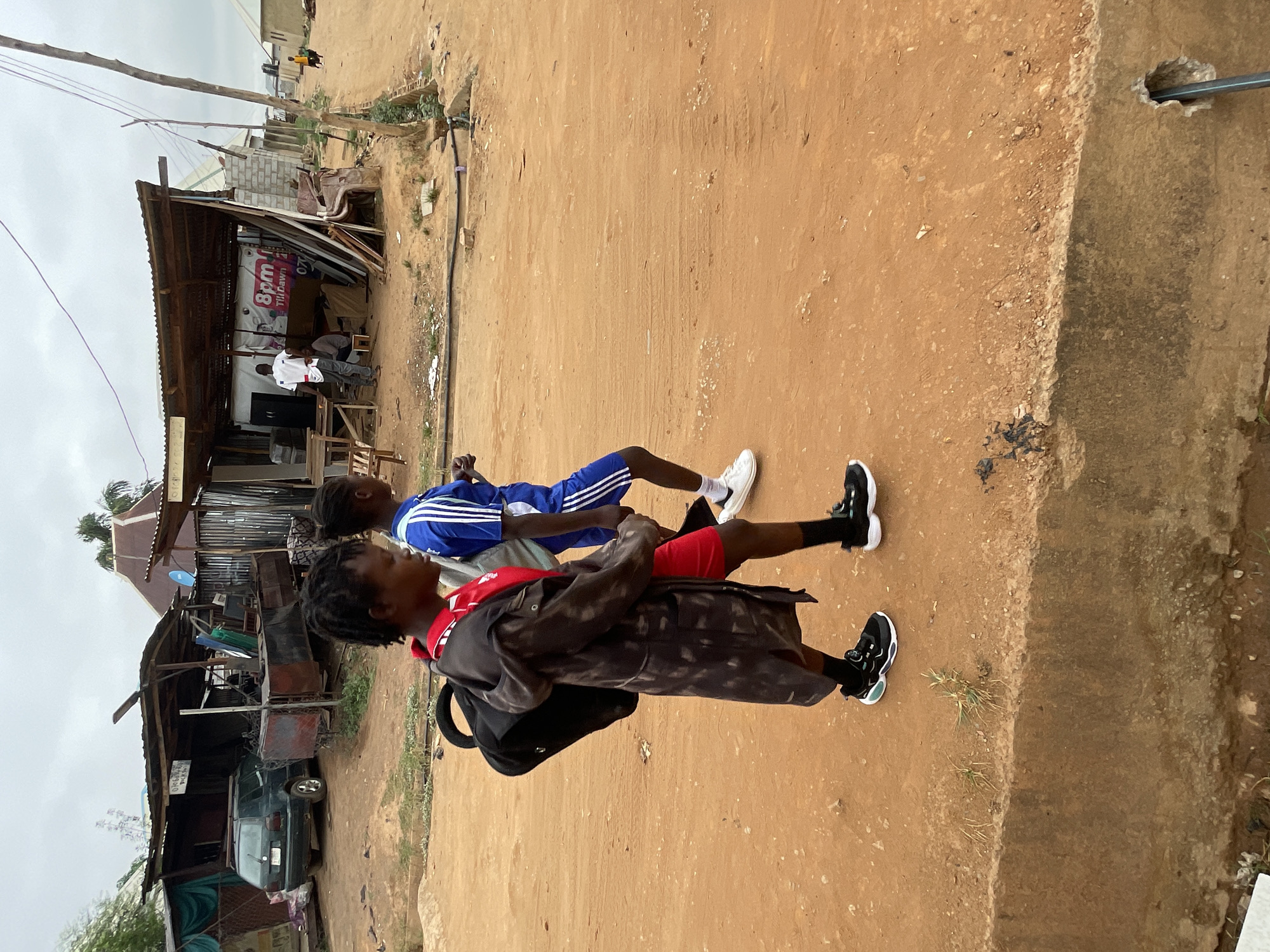 Two young men are walking along a dirt road in Karmajiji, Nigeria. The man in front wears a dark coat, a red shirt, and black shorts. He carries a black backpack. The second man wears a blue and white track suit. Both men wear athletic shoes. The road is bordered by a concrete curb on the right. Dilapidated structures and vehicles are visible on the left side of the road. Overhead wires are present. The sky is overcast.