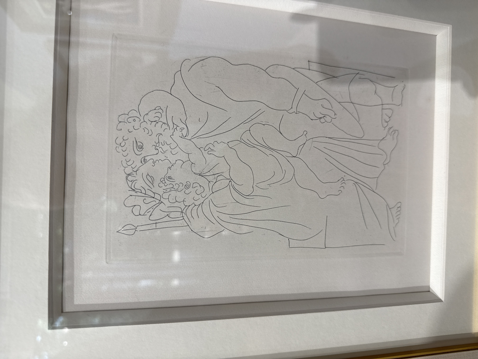 A framed line drawing depicts two figures in an embrace. The figure on the left is smaller, with curly hair, wearing a robe and a crown adorned with foliage and a flame-like object. They are seated and lean towards the larger figure on the right. The larger figure is muscular with curly hair and a beard, wearing a draped garment. Their faces are close, suggesting a kiss or intimate moment. The artwork is presented in a white mat and a silver frame. The background appears to be a white wall.