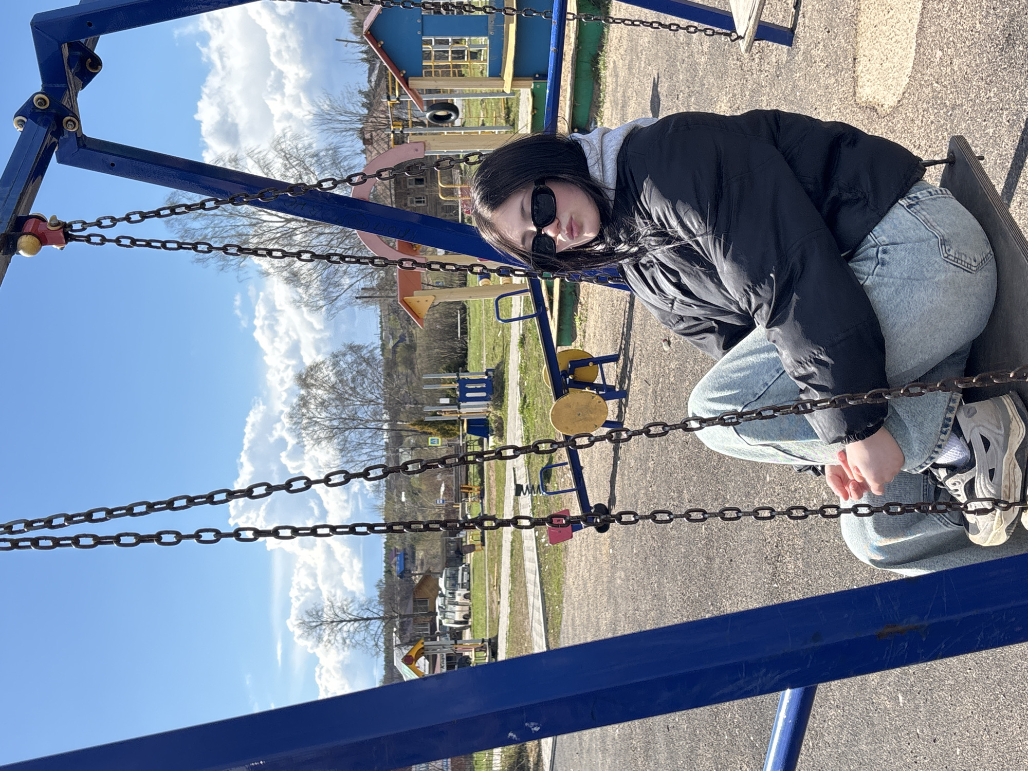 A young woman is captured in a vertical, outdoor full-shot, sitting on a swing at a playground. She is dressed in a black puffy jacket, a grey hoodie, light blue jeans, and grey and white sneakers, with dark sunglasses obscuring her eyes. Her dark hair frames her face, and her lips are slightly pursed, conveying a reserved expression.
She is seated on a dark swing seat, suspended by thick metal chains from a prominent blue metal swing frame that dominates the left foreground. The background reveals a bright, sunny day under a clear blue sky with fluffy white clouds. Scattered trees with bare branches suggest an early spring or late autumn setting.
Behind the swing, other colorful playground equipment, including a small house-like structure and a yellow and red seesaw, are visible. The ground beneath the playground appears to be paved or packed earth. In the mid-distance, residential buildings and what look like parked vehicles are present, along with a path or road. The overall setting appears to be a community playground in a semi-rural or residential area, consistent with the given location of Фоки, Russia. Some faint, unreadable Cyrillic text can be seen on a distant yellow sign.
