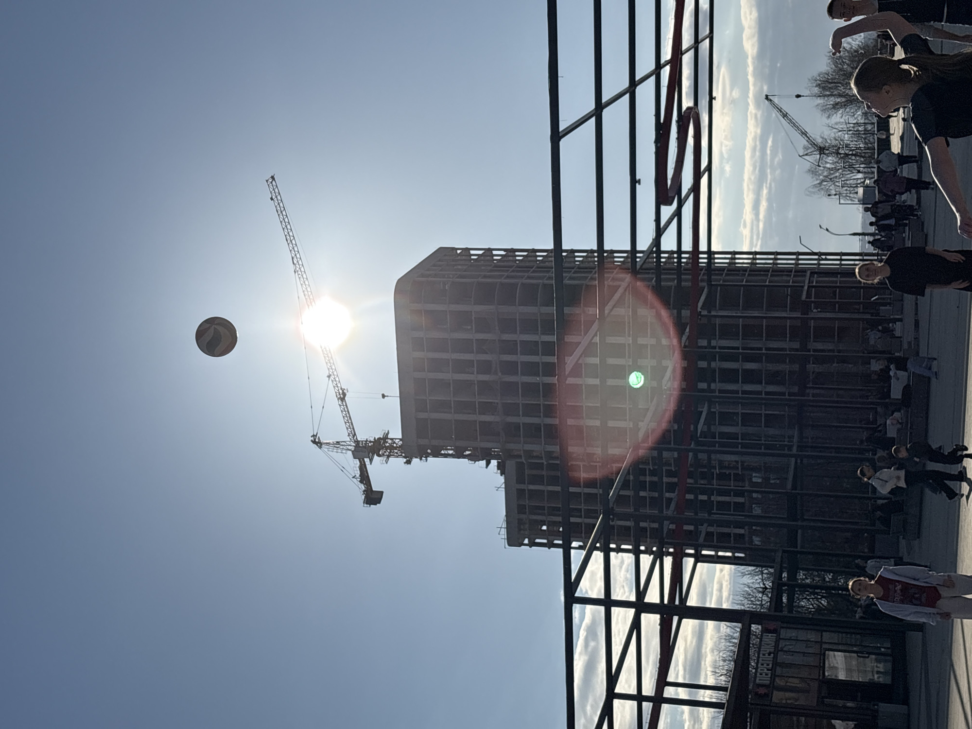 The photograph shows a partially constructed high-rise building in Izhevsk, Russia, with two cranes in operation around it. A ball floats in the sky next to the sun. In the foreground, a metal structure partially obscures the view. People are seen walking along a path near the building. A small kiosk is also visible in the lower right corner of the frame.