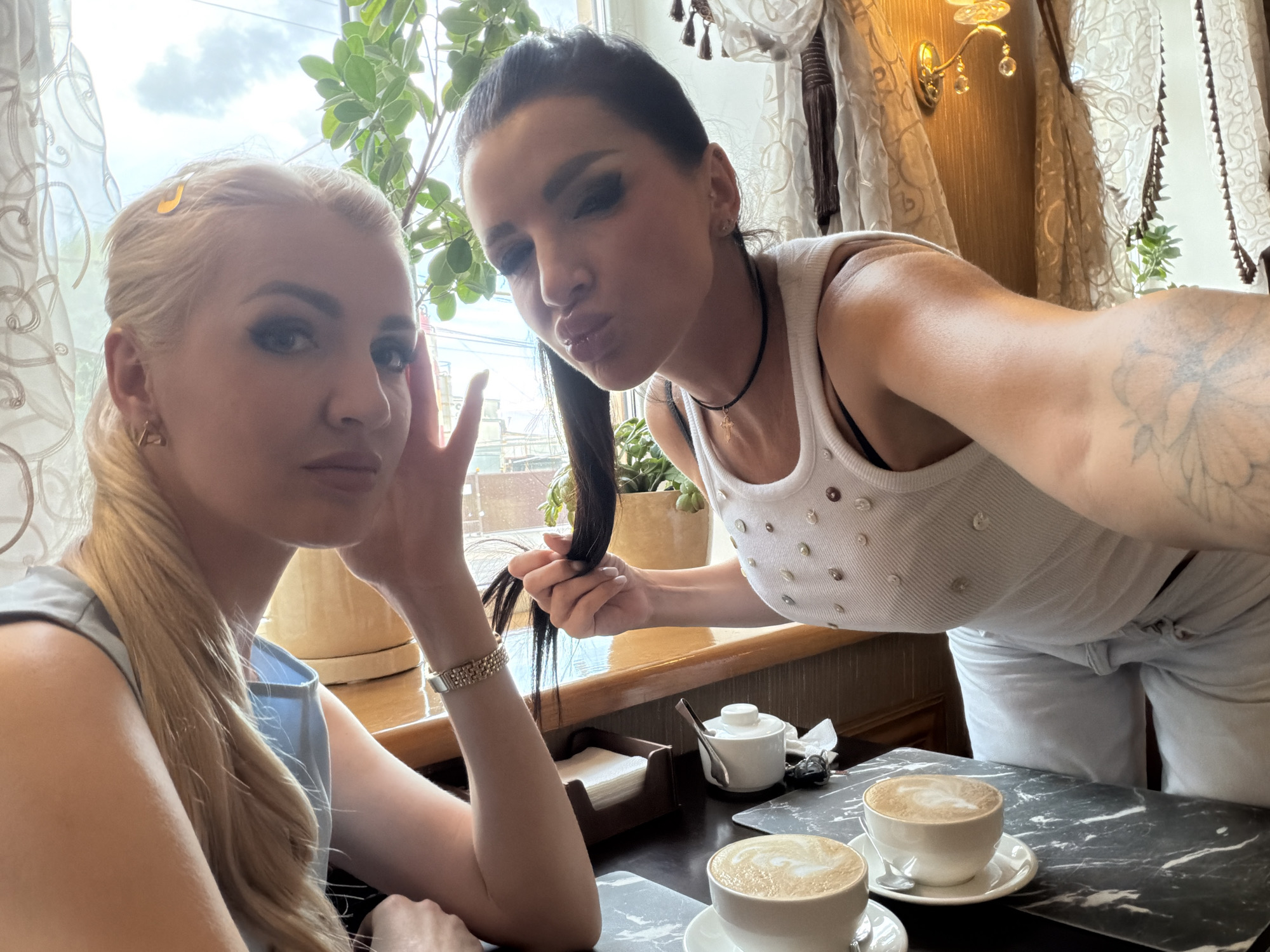 Two women are taking a selfie at a cafe table in Novaya Usman', Russia. The woman on the left, with blonde hair in a ponytail, wears a light blue top and a watch. The woman on the right, with dark hair, wears a white tank top with rhinestones and a necklace. She is leaning towards the camera and puckering her lips. In front of them are two cups of coffee on a table with a marble-like pattern. Behind them, a window with sheer curtains reveals greenery and a cloudy sky. A wall sconce with crystals is visible on the right wall.